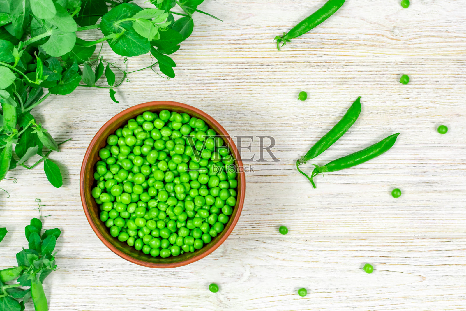 碗里装着新鲜的绿豌豆,背景是嫩叶,白色木桌上放着绿豌豆的小枝。从以上观点。适当的营养,维生素。照片摄影图片