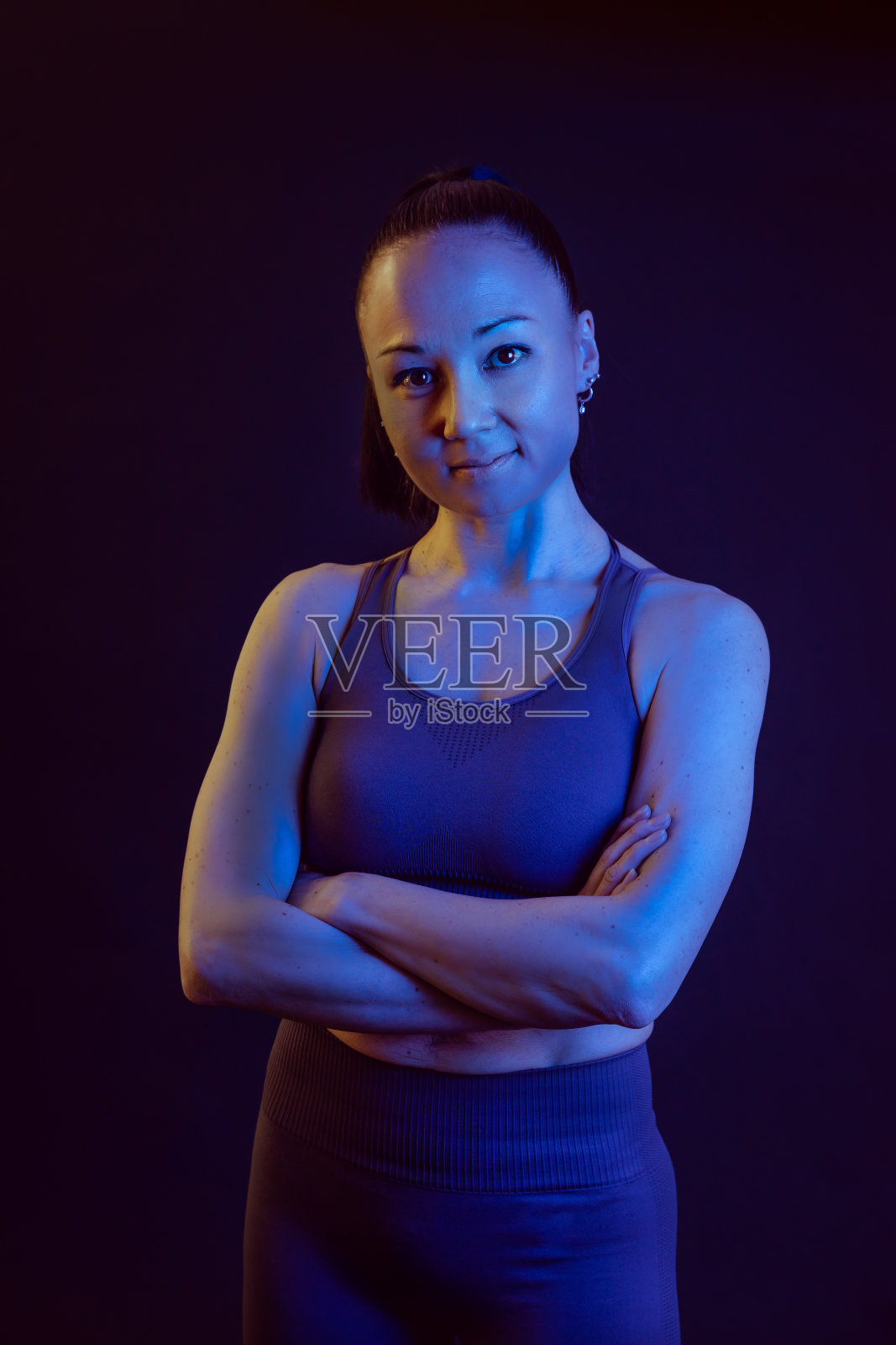 垂直工作室的时尚肖像,一个亚洲女人,健身教练,穿着运动服,黄色和蓝色霓虹灯。这名女子双手交叉,对着镜头微笑,表情坚定而自信。照片摄影图片