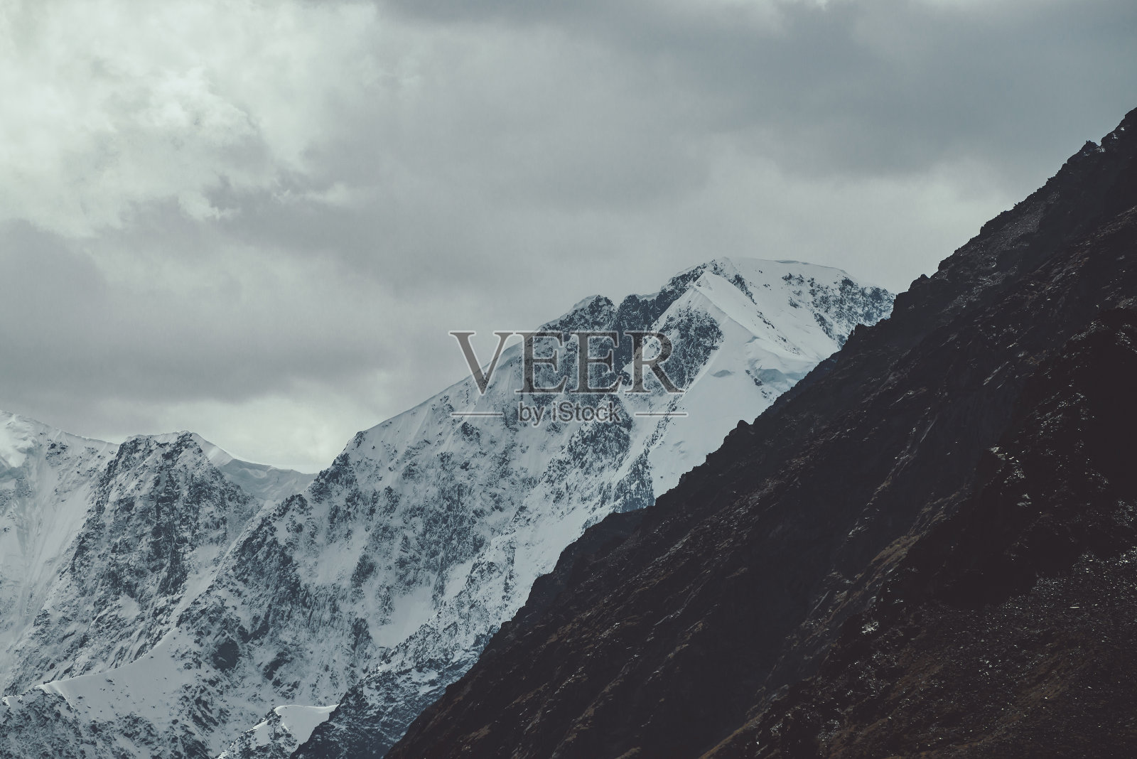 大气景观与巨大的雪山墙在云。阴沉的山景与高积雪覆盖的山顶下多云的天空褪色的色调。雪峰和暗色岩石。照片摄影图片