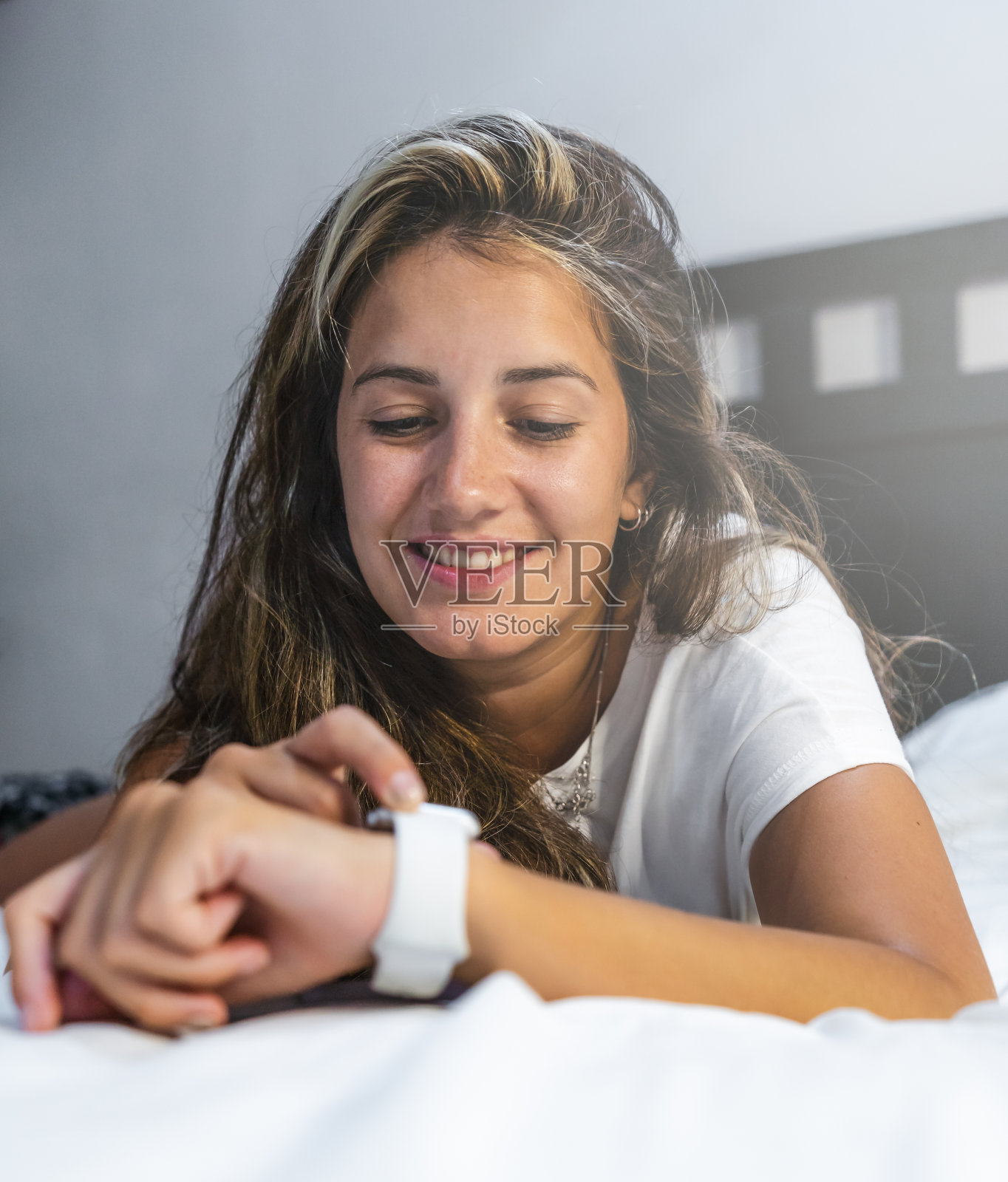 年轻的西班牙妇女躺在床上,穿着睡衣,用她的智能手表微笑照片摄影图片