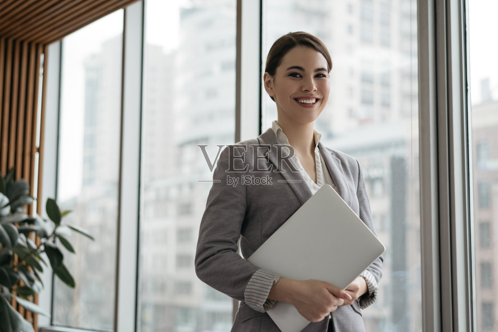 自信的女商人拿着笔记本电脑,站在办公室靠窗的地方。成功经理的肖像,穿着正装看着相机和微笑照片摄影图片