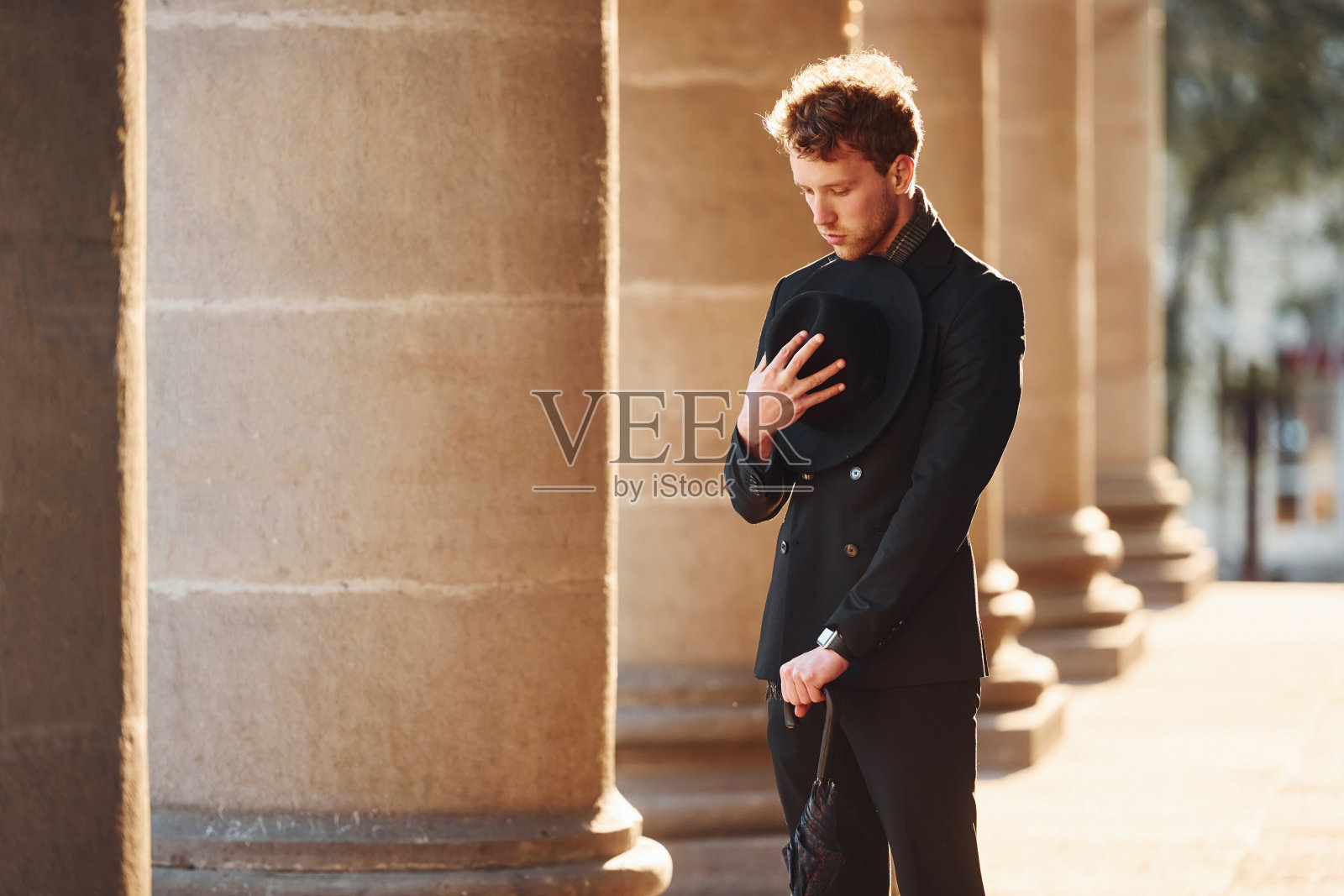 优雅的年轻人戴着黑帽子,穿着黑衣服,手里拿着雨伞在城外照片摄影图片