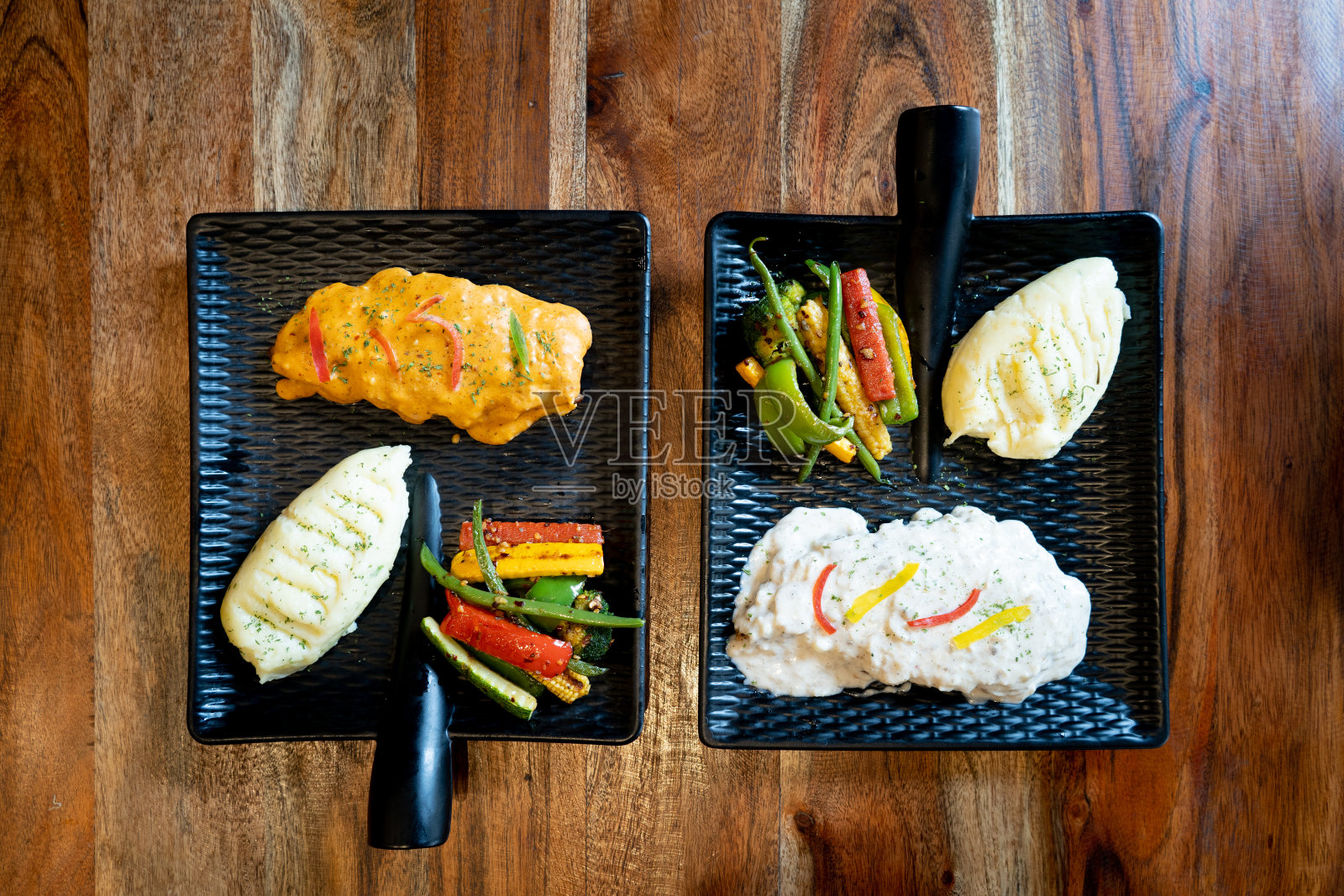 平面照片显示了两个美观的黑色方形带把手的盘子放在木桌上,里面装满了白酱意大利面和红酱土豆泥和辣椒蔬菜沙拉照片摄影图片