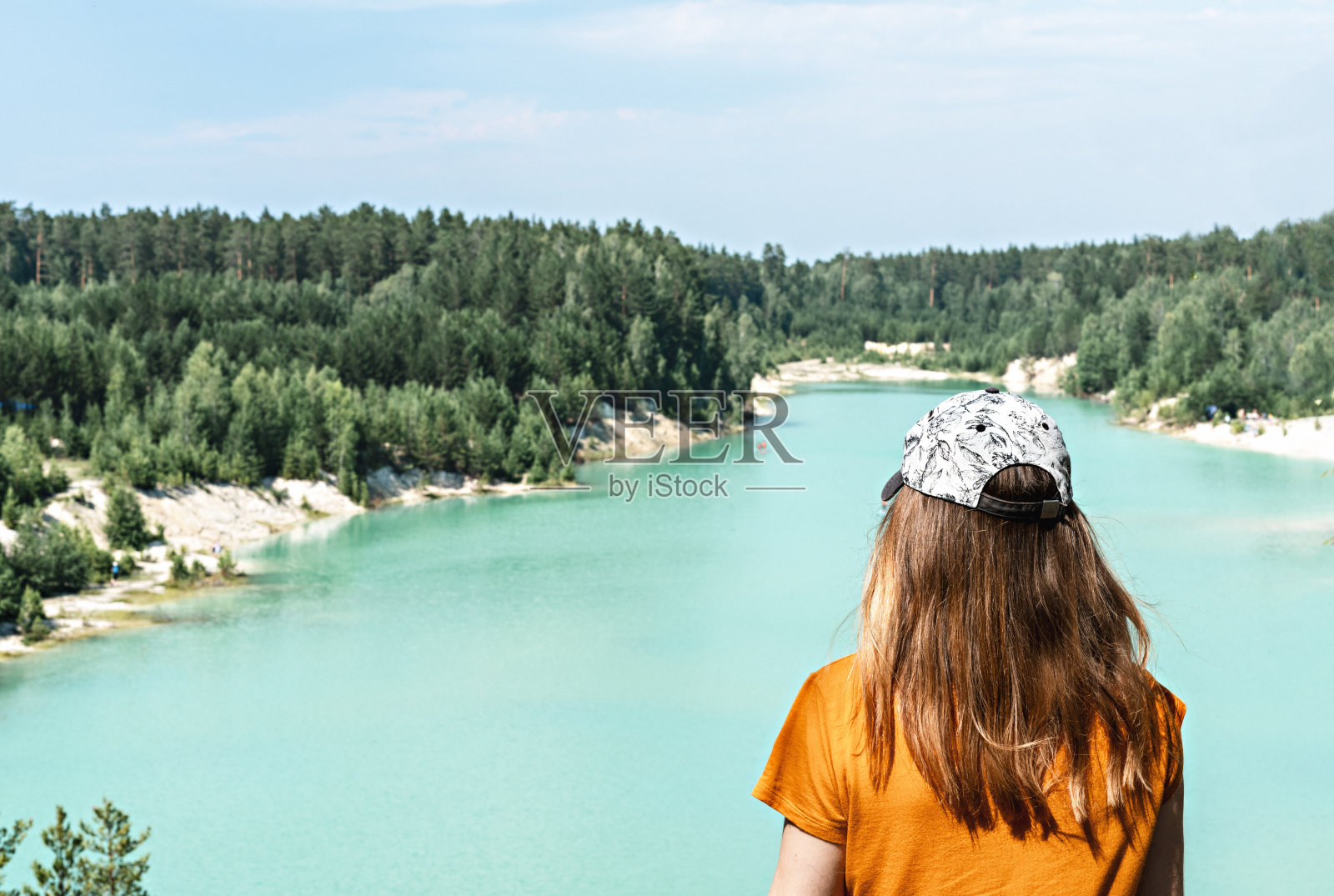 后视图的年轻的金发女人旅行者在帽子和姜色t恤站在一个蓝色的湖或河流和针叶林悬崖上,旅行美丽的风景照片摄影图片