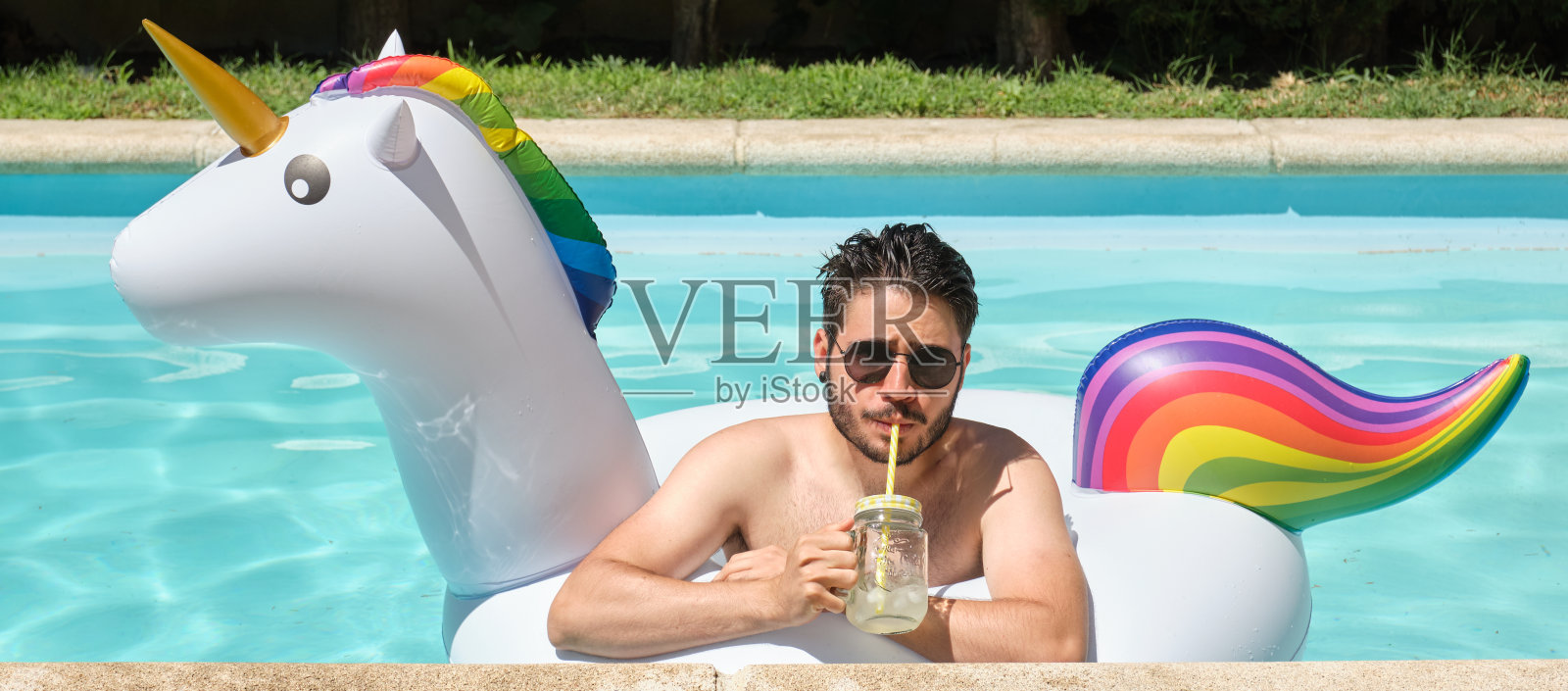 一个戴着墨镜的年轻人在游泳池里的独角兽充气圈里喝柠檬水的全景图。夏天的概念。照片摄影图片