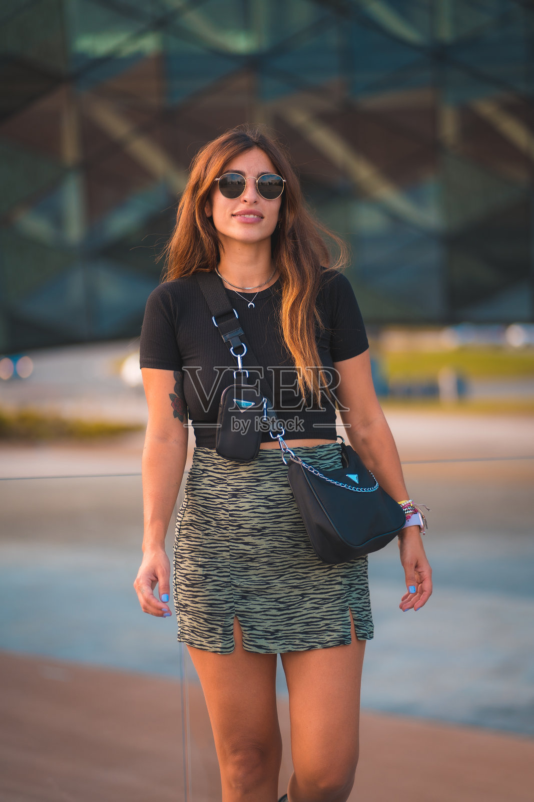 年轻漂亮的黑发白种女商人在后台办公大楼外。穿着绿色裙子,戴着太阳镜走路照片摄影图片