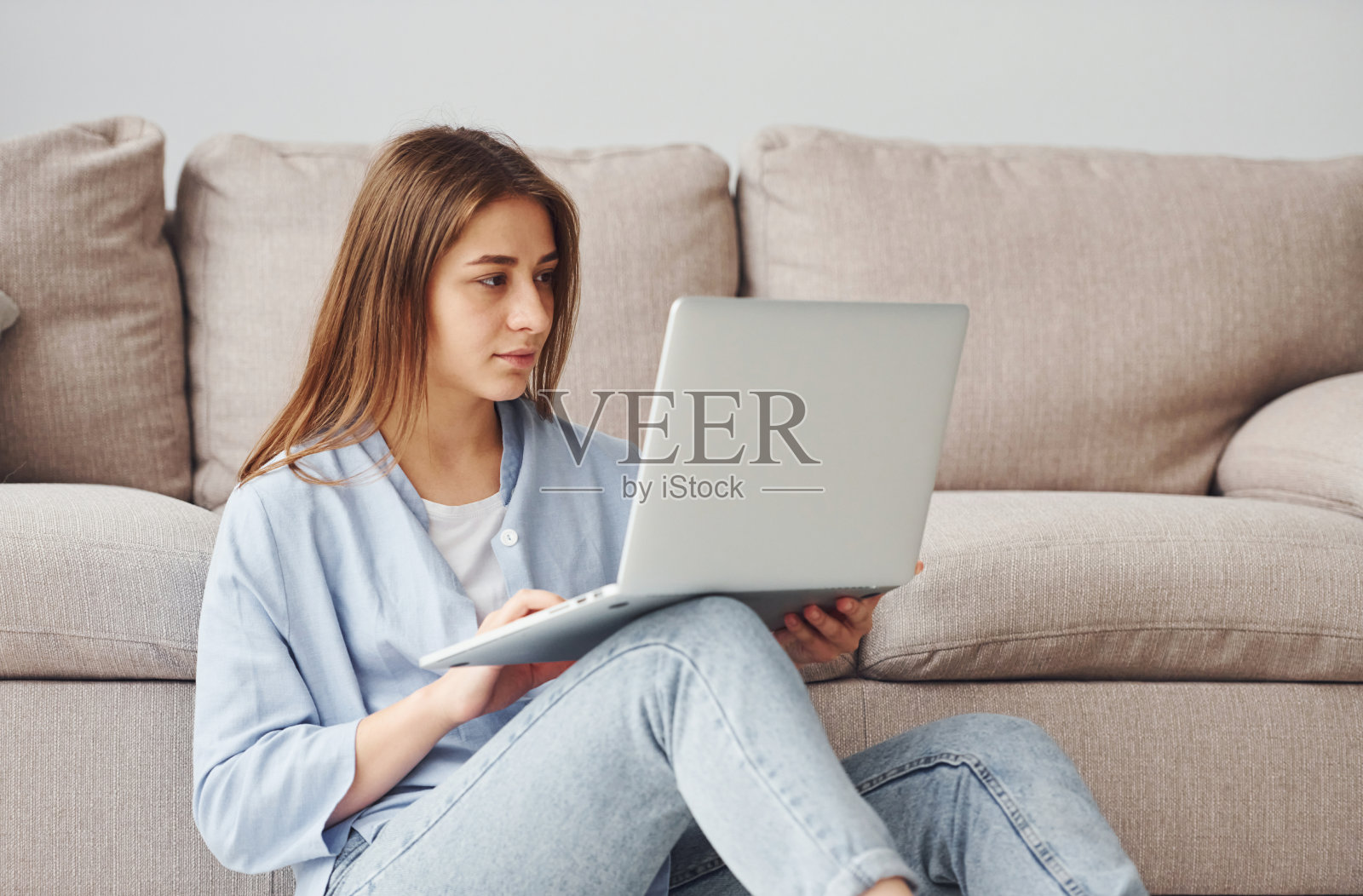 穿着牛仔裤和蓝色衬衫的漂亮年轻女子坐在家里的地板上,手提电脑照片摄影图片