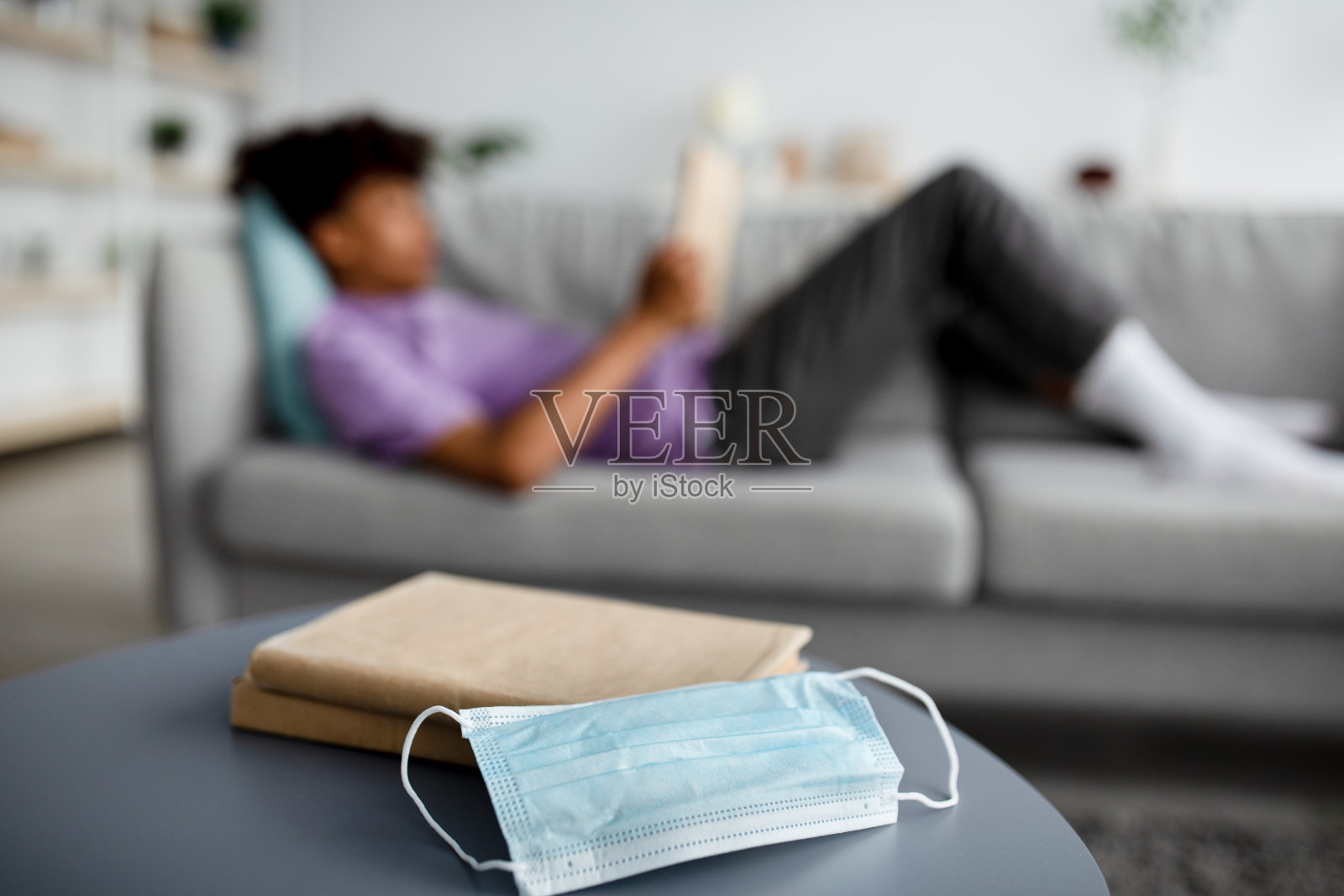 模糊的黑人少年在沙发上看书,准备考试,选择性地把医用口罩和课本放在桌子上照片摄影图片