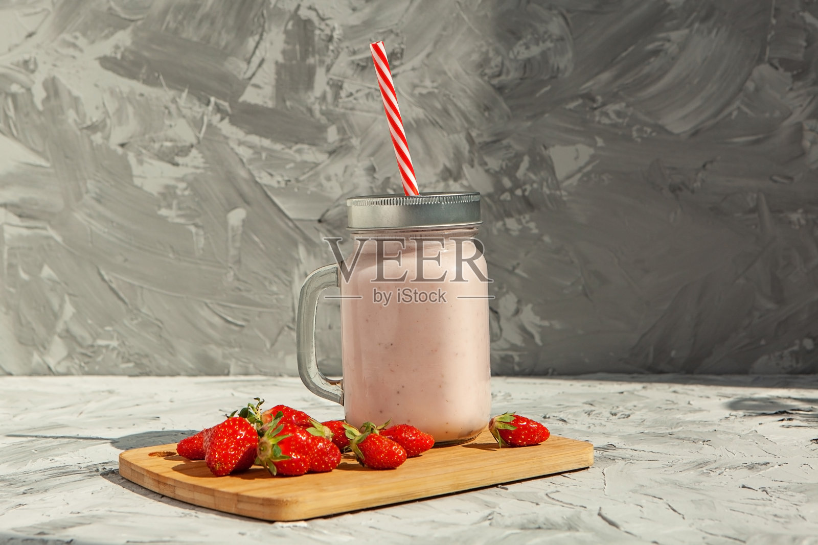 草莓冰沙,装在玻璃罐里,有红色条纹管。木板上放着草莓,旁边放着草莓奶昔。健康食品的概念。在浅混凝土背景上照片摄影图片