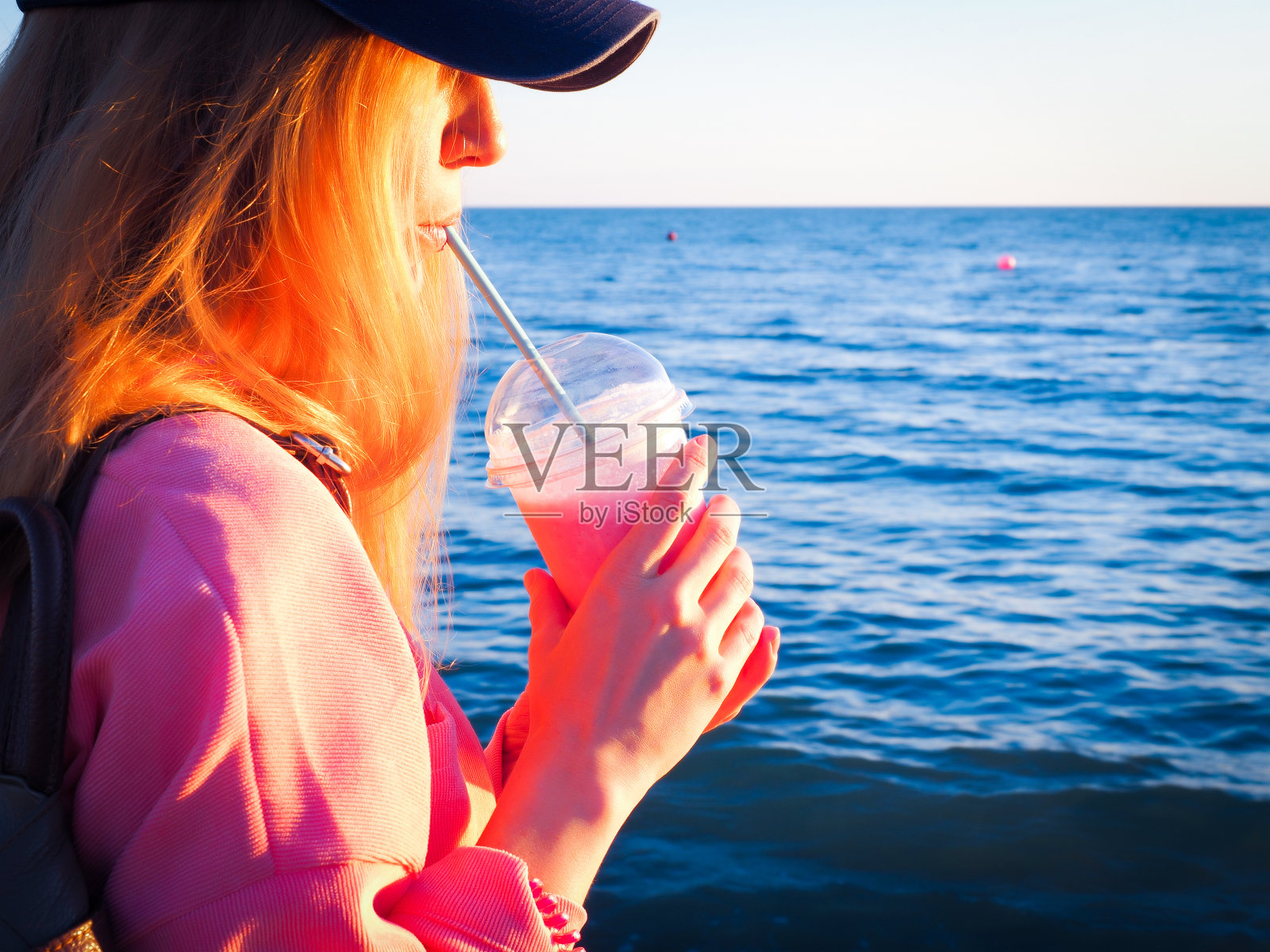 女孩从一个以大海为背景的管子里喝着粉红色的奶昔照片摄影图片