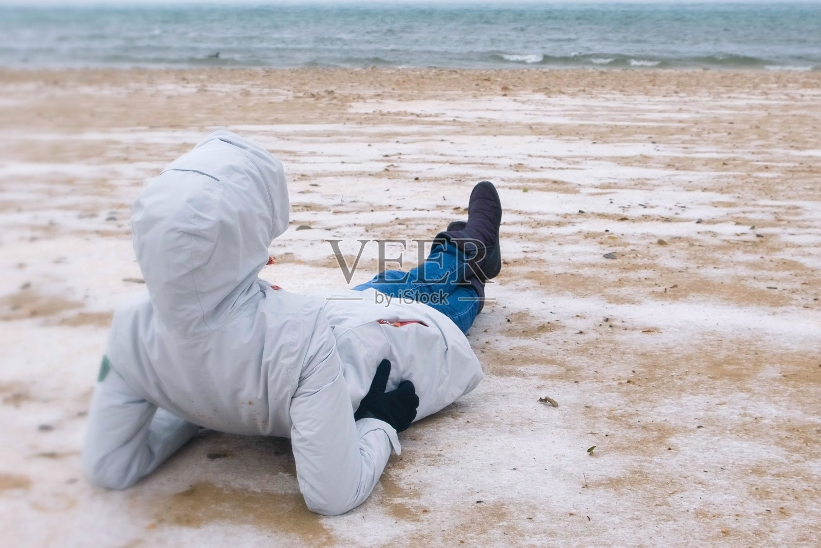 冬天,一名女游客躺在海边白雪皑皑的沙滩上。穿着夹克和牛仔裤的女人,背影。照片摄影图片