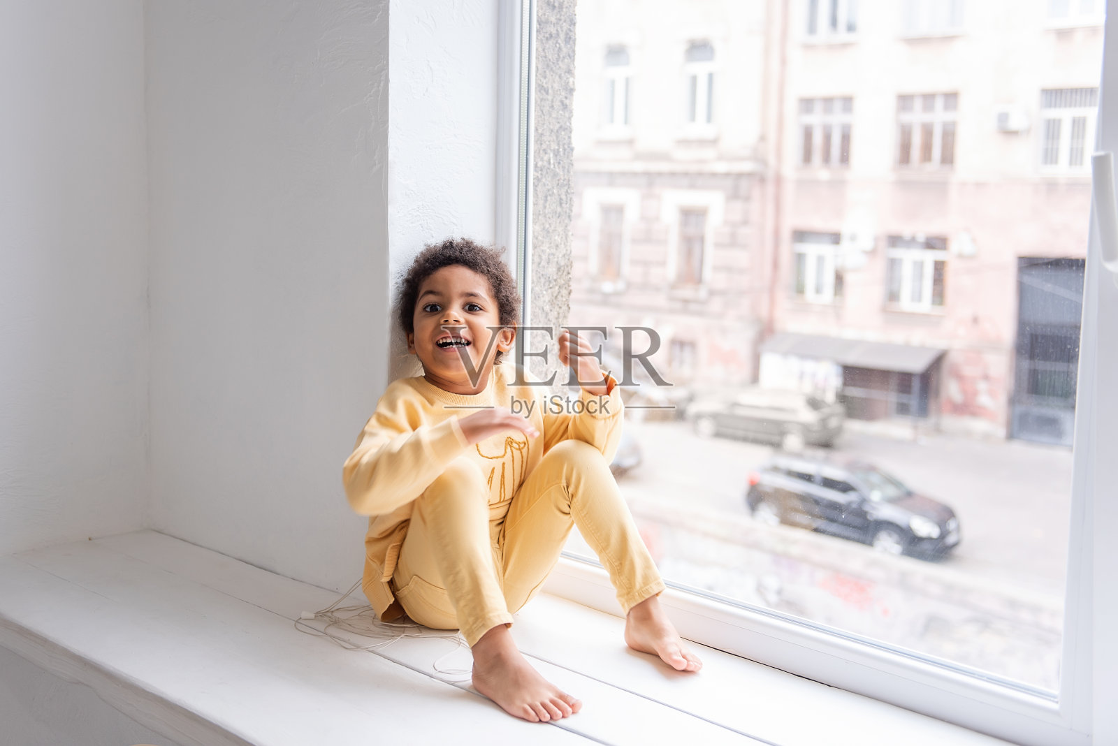 一个穿着黄色睡衣的非裔美国小男孩赤着脚坐在一扇白色大窗户旁边的宽窗台上。这孩子玩得很开心,又笑又闹。快乐的生活方式童年在家里照片摄影图片