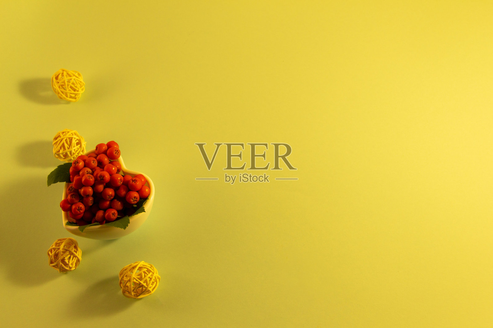 花楸树枝在一个心形花瓶上,黄色的背景,橙色的花楸浆果和绿色的叶子。圆形装饰球,文字背景照片摄影图片