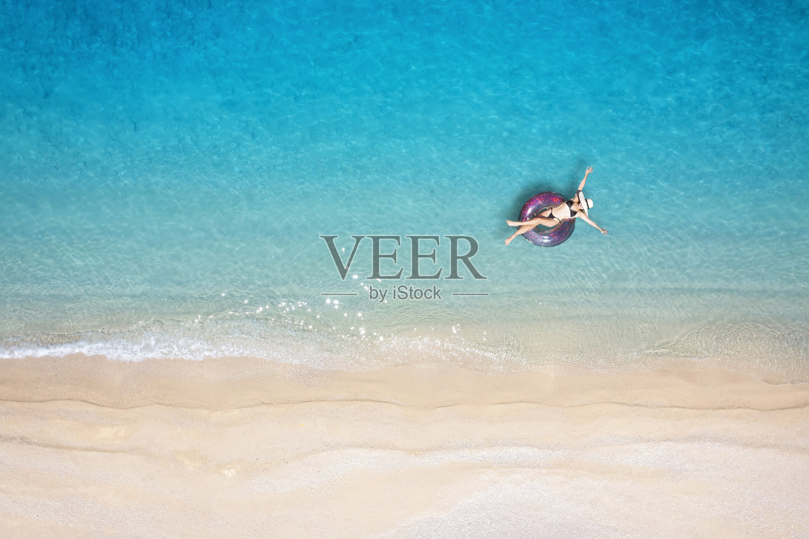 一名身穿比基尼的女子在一个甜甜圈形状的充气气球上漂浮,漂浮在碧绿、清澈的大海上照片摄影图片