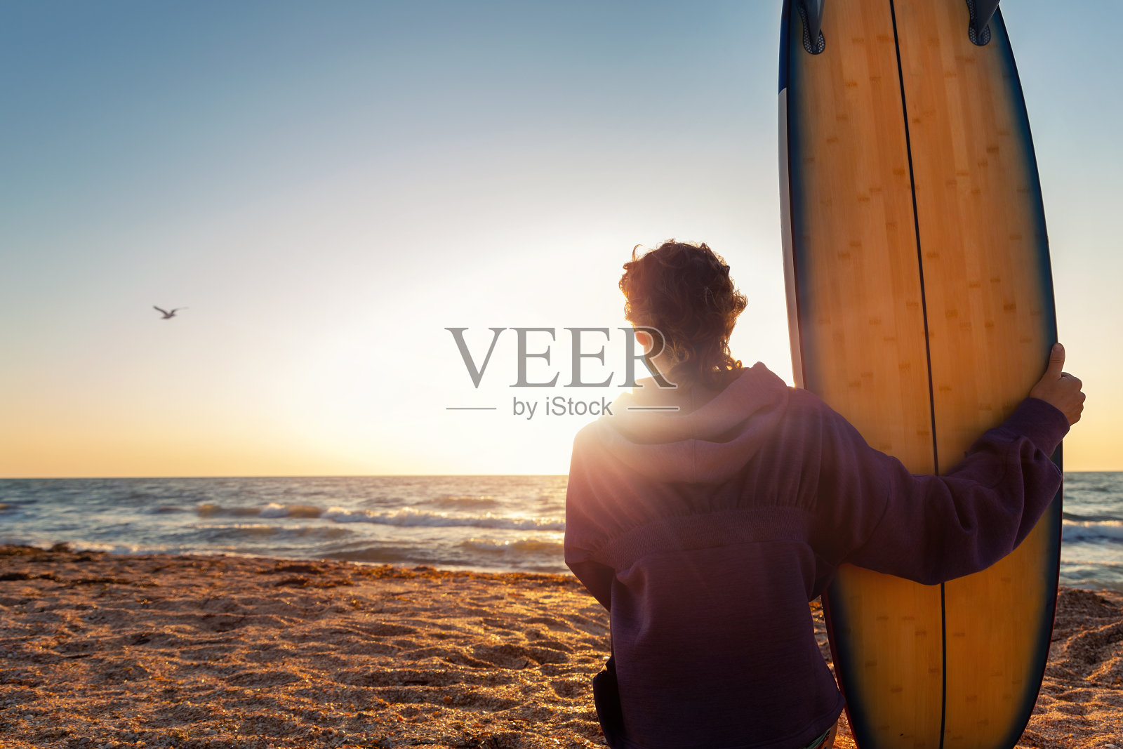 背视图年轻的成年苗条运动女性冲浪女孩冲浪板坐在沙滩上的海洋海岸波浪反对温暖的日出或日落的太阳。运动,健康,无忧无虑,自由的生活方式,度假的概念照片摄影图片