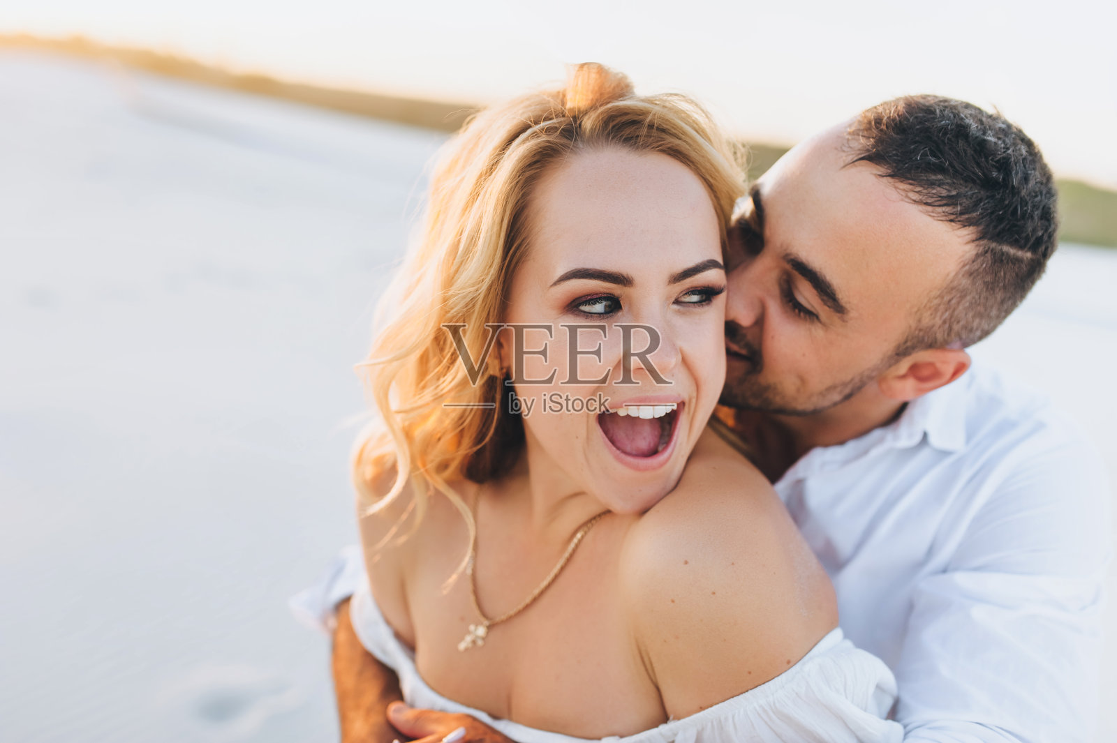 在夕阳的背景下,一个皮肤黝黑的年轻白人男子亲吻了一个美丽有趣的大嘴巴金发女人。沙漠,沙滩,休息和放松。蜜月的概念。照片摄影图片