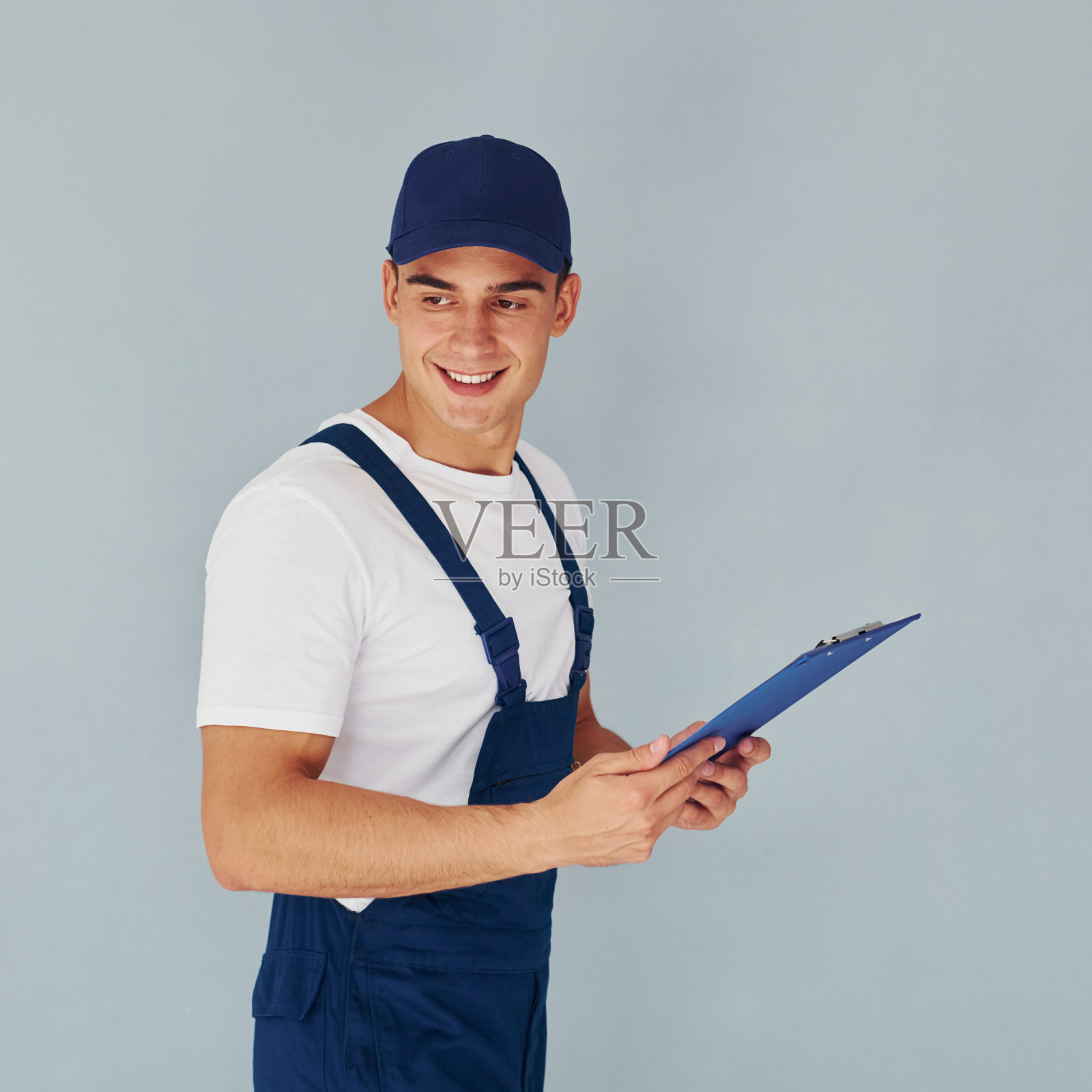 戴上帽子,用记事本。身穿蓝色制服的男员工站在白色背景下的工作室内照片摄影图片
