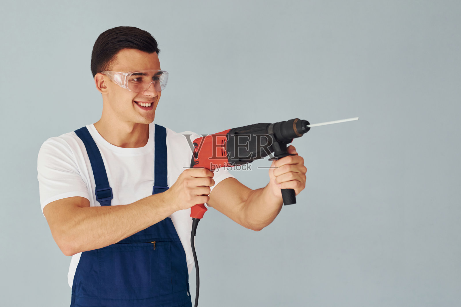 戴着防护眼镜,手里拿着钻头。身穿蓝色制服的男员工站在白色背景下的工作室内照片摄影图片