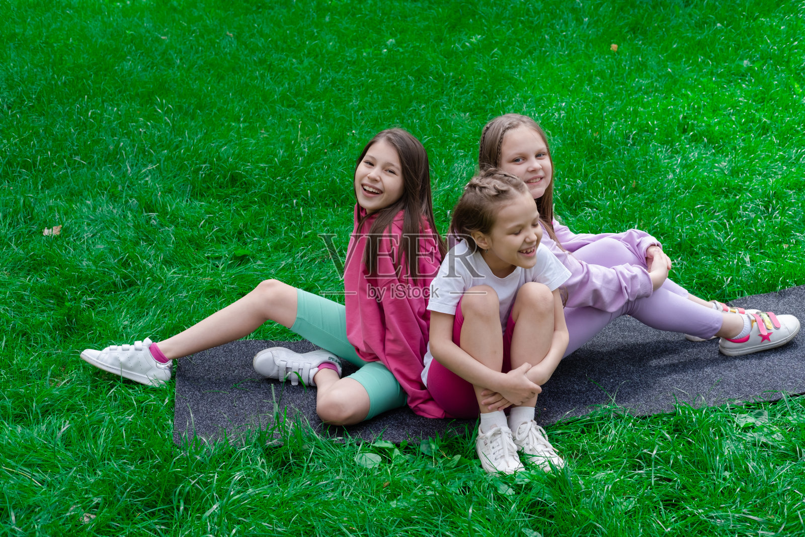 三个美丽快乐的十几岁的少女,穿着五颜六色的衣服,坐在公园的绿草地上。夏季。快乐的孩子。假期,暑假。姐妹,最好的朋友,闺蜜,姐妹情谊照片摄影图片