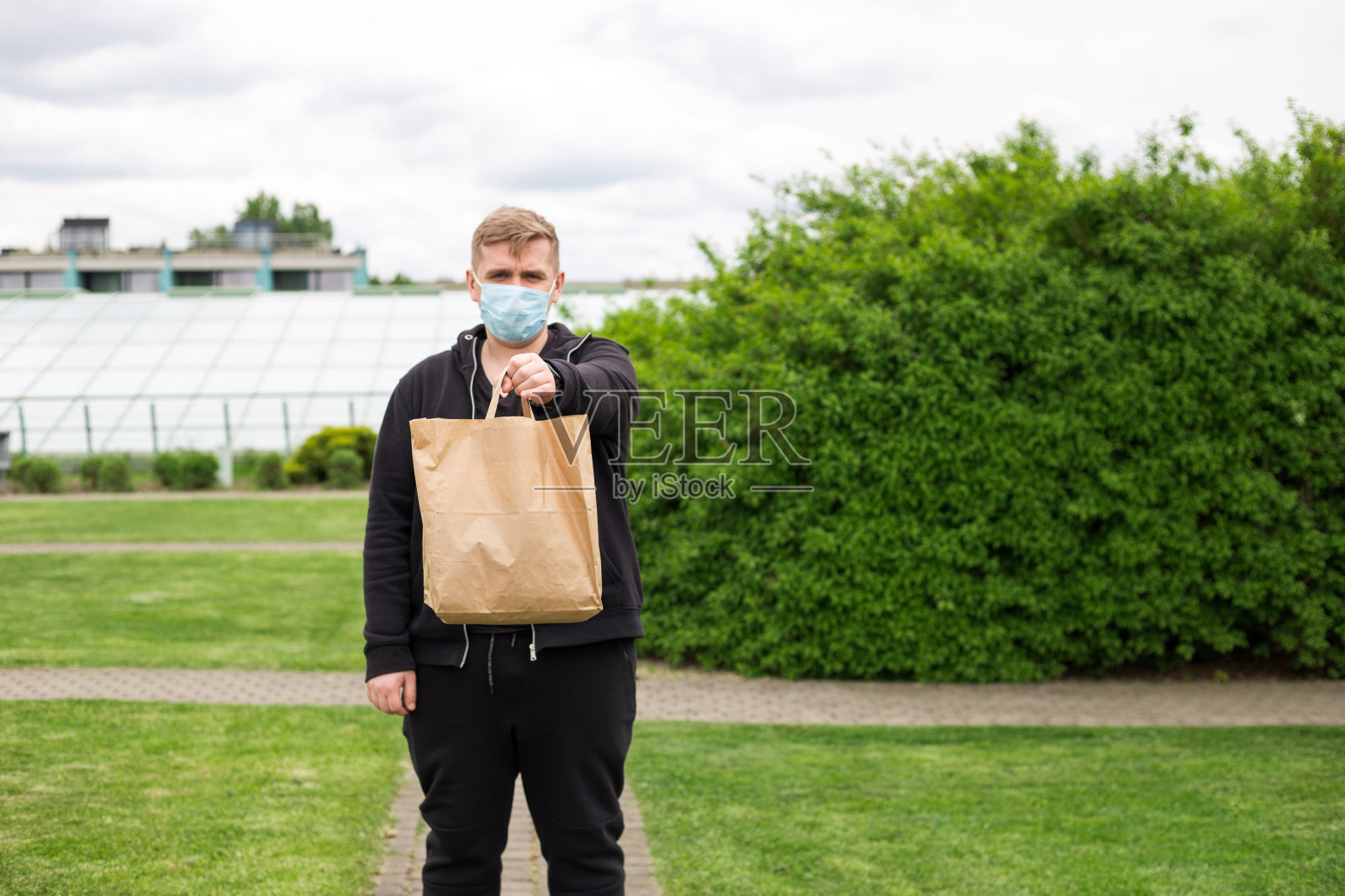 在大自然的绿色背景下,男子手戴口罩,手拿外卖纸袋。在任何天气条件下24小时送货给客户。照片摄影图片