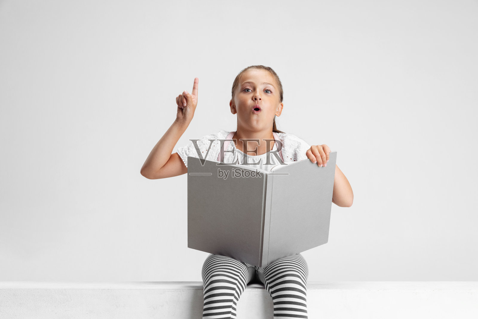 可爱的白人女孩的肖像,青少年坐在和阅读大的书,杂志孤立在灰色的工作室背景。童年,教育,情感观念。照片摄影图片