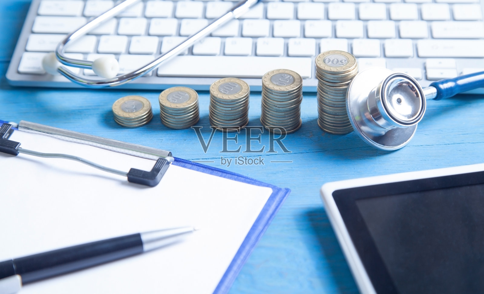 听诊器、平板电脑、电脑键盘和蓝色背景上的硬币。照片摄影图片