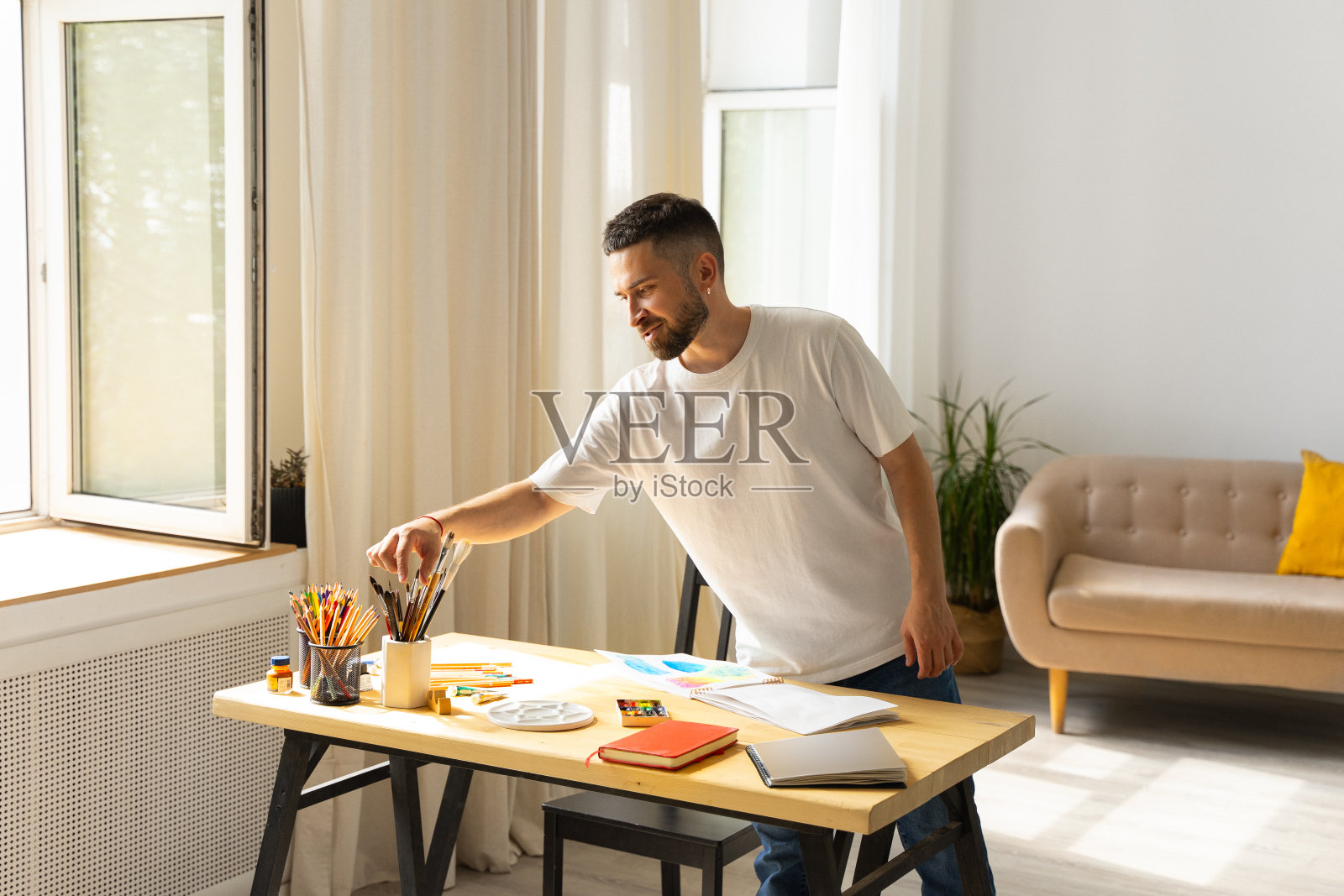 可爱的白种人男性艺术家,深色头发和胡子,穿着白色t恤,拿着刷子从石膏玻璃和木桌上的彩色铅笔开始画画。现代的房间照片摄影图片