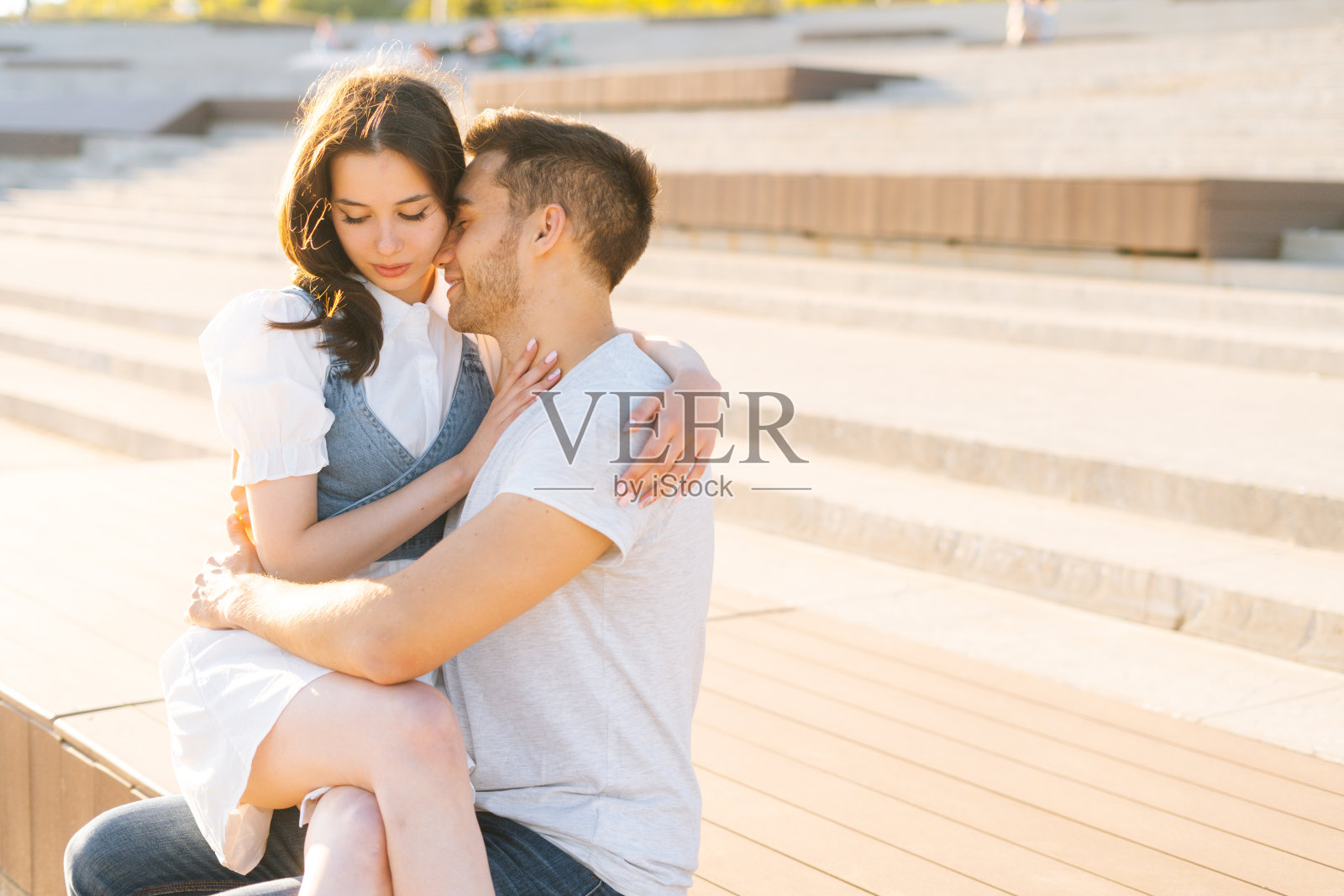 在夏日阳光明媚的日子里,一对美丽的年轻夫妇在城市公园休息,坐在长椅上拥抱。照片摄影图片