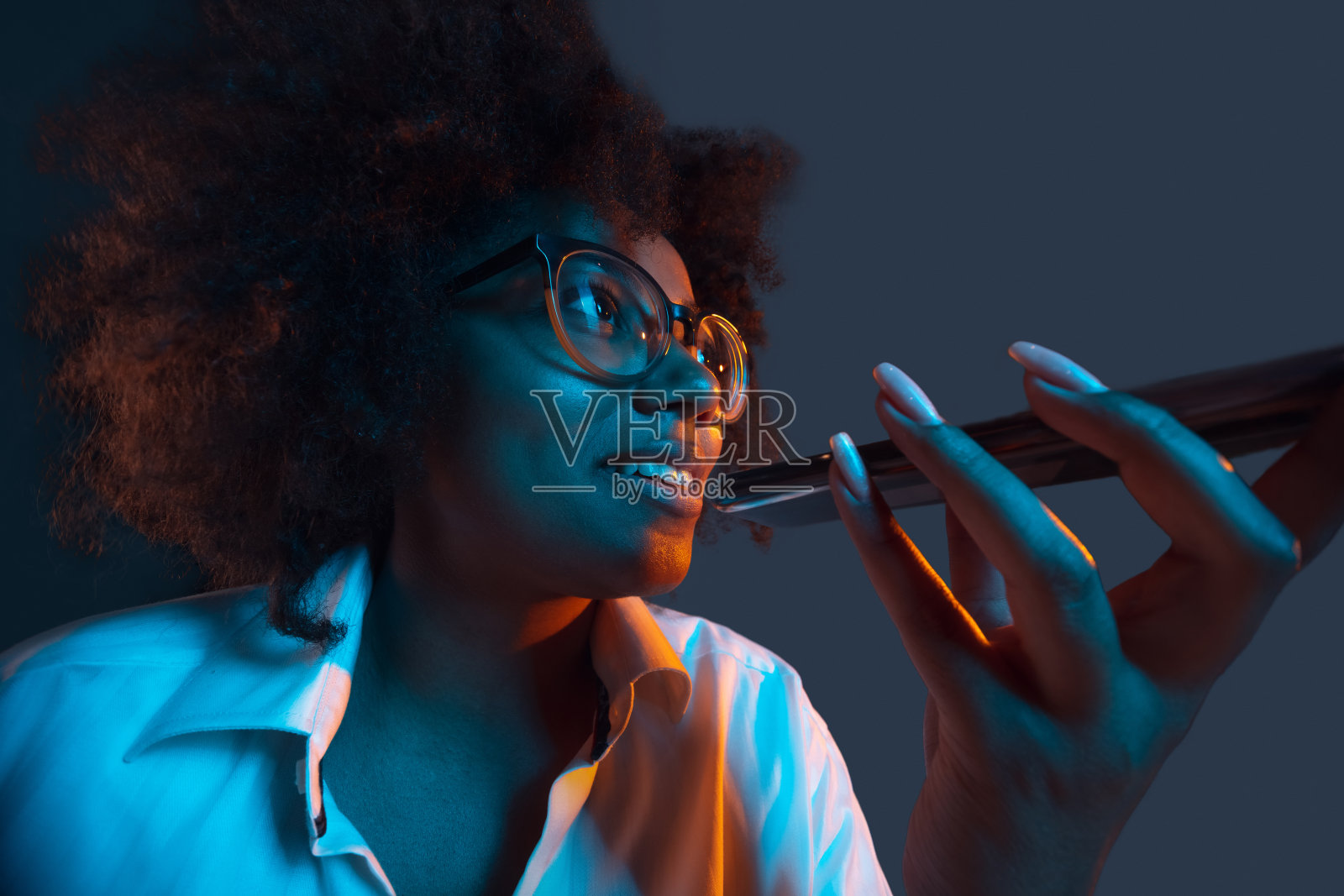 非洲年轻女孩的肖像在白色衬衫使用手机,小工具孤立在深蓝色的工作室背景。人类情感概念,面部表情,青春,销售,广告。照片摄影图片