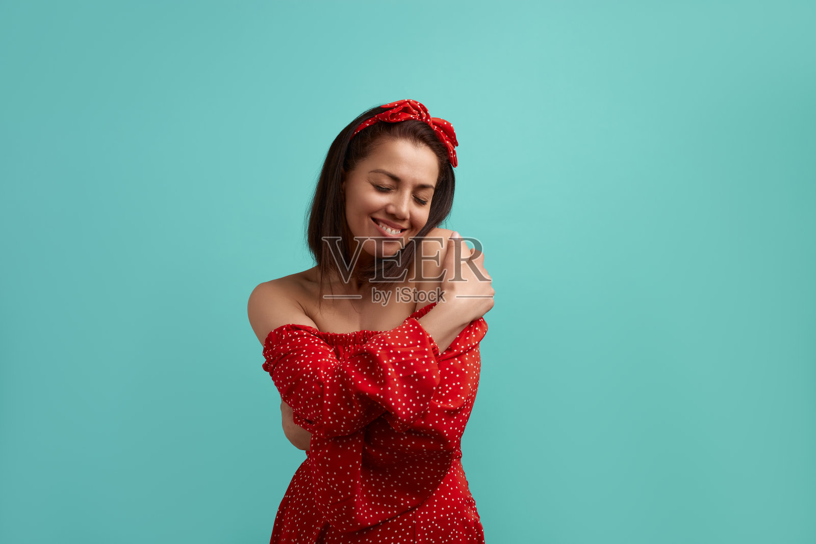 快乐的白种人女性,红裙裸肩带发带的黑发回忆浪漫的记忆,拥抱自己,抚肩,想你,孤立在绿松石的背景上。照片摄影图片