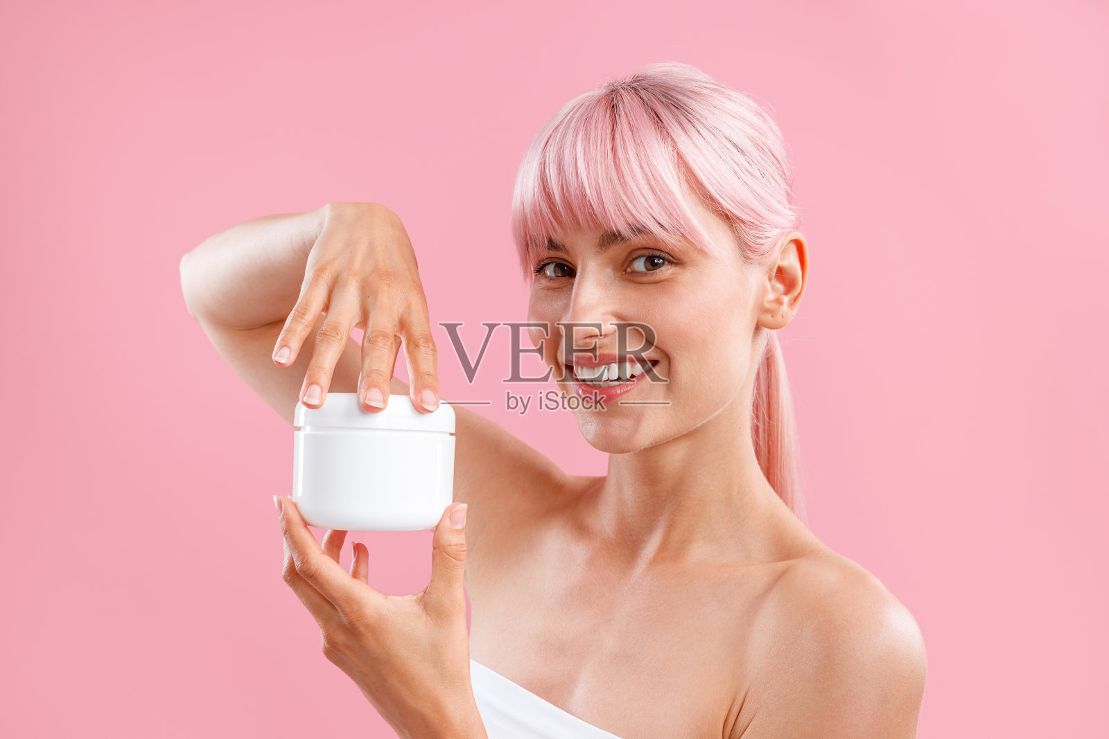 一名年轻女子的肖像,对着镜头微笑,在粉红色的背景下,拿着一个白色的罐子,里面装着润肤露照片摄影图片