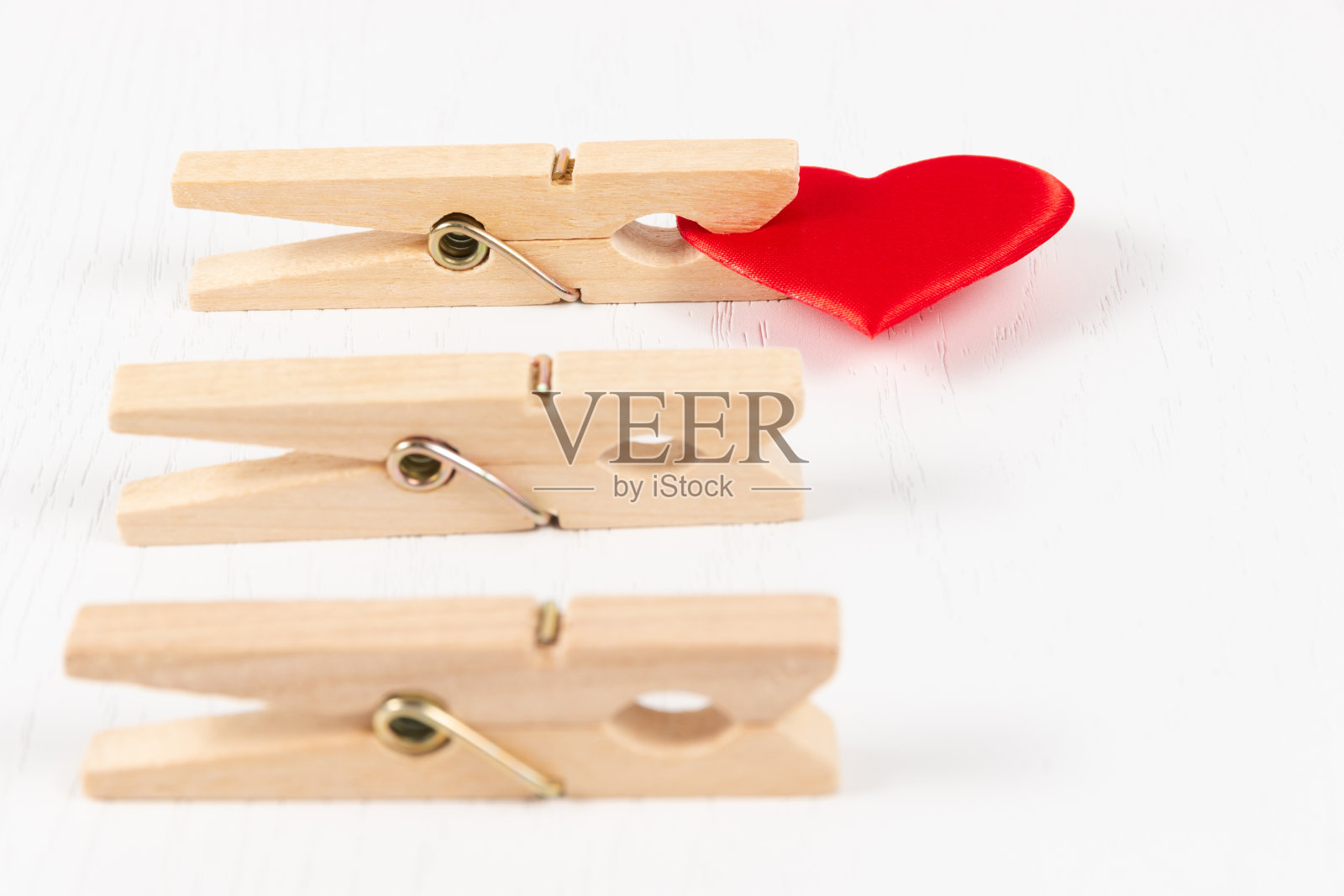白色木桌背景上,三个带有金属弹簧的木衣夹夹着一颗红心。特写镜头照片摄影图片