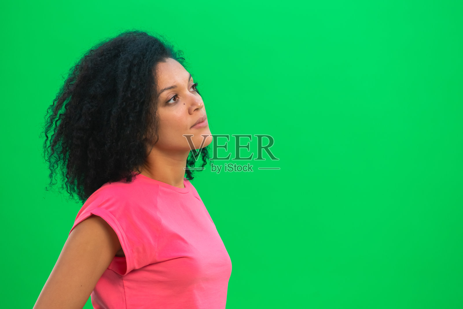 年轻的非裔美国女性的肖像考虑和选择的东西。一名身穿粉色t恤、一头卷发的黑人女子在演播室的绿色屏幕上摆姿势。近距离照片摄影图片