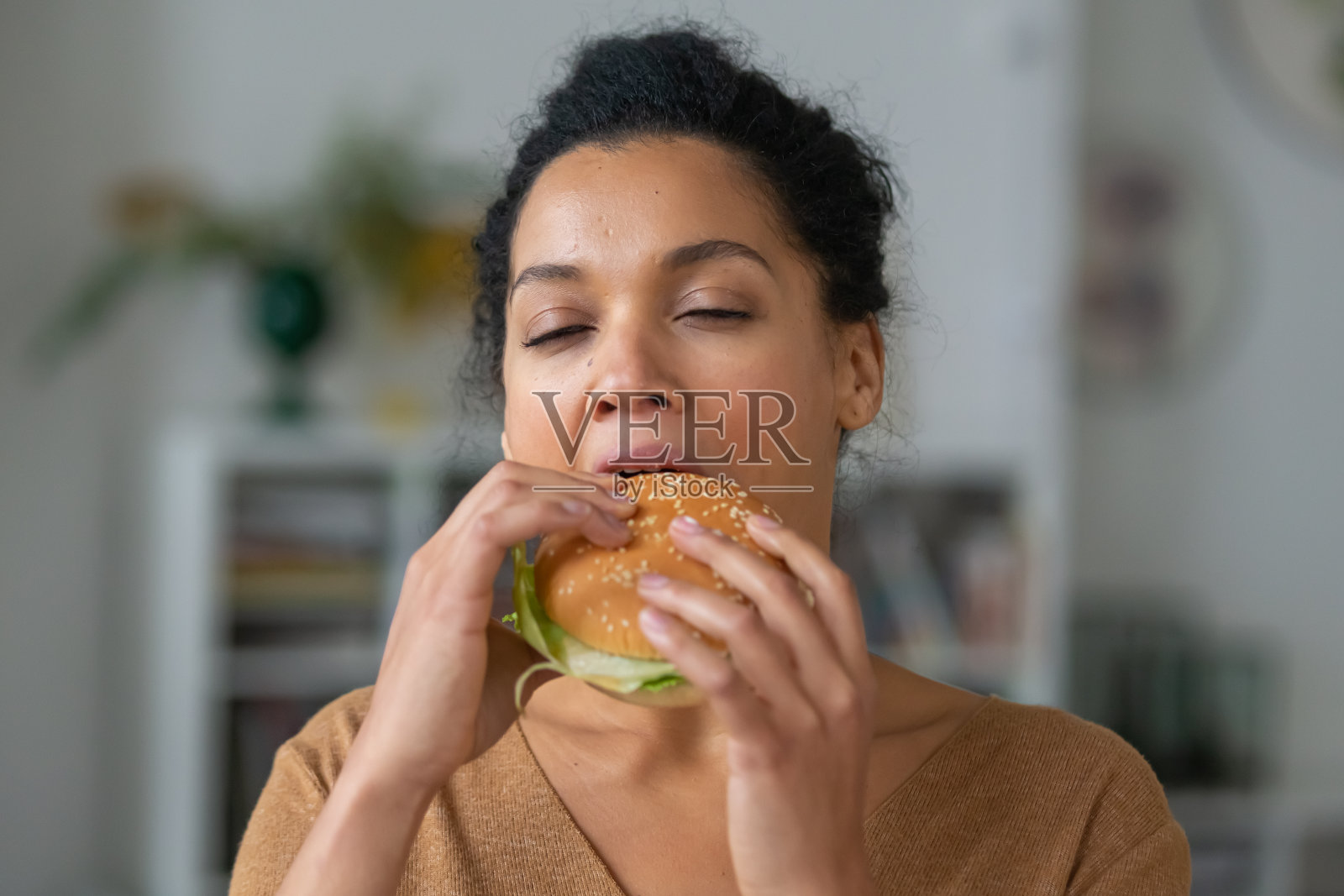 漂亮的非裔美国女人的肖像,胃口大开,喜欢咬汉堡。年轻的混血女性吃着快餐,在光线模糊的背景下摆着姿势。近距离照片摄影图片