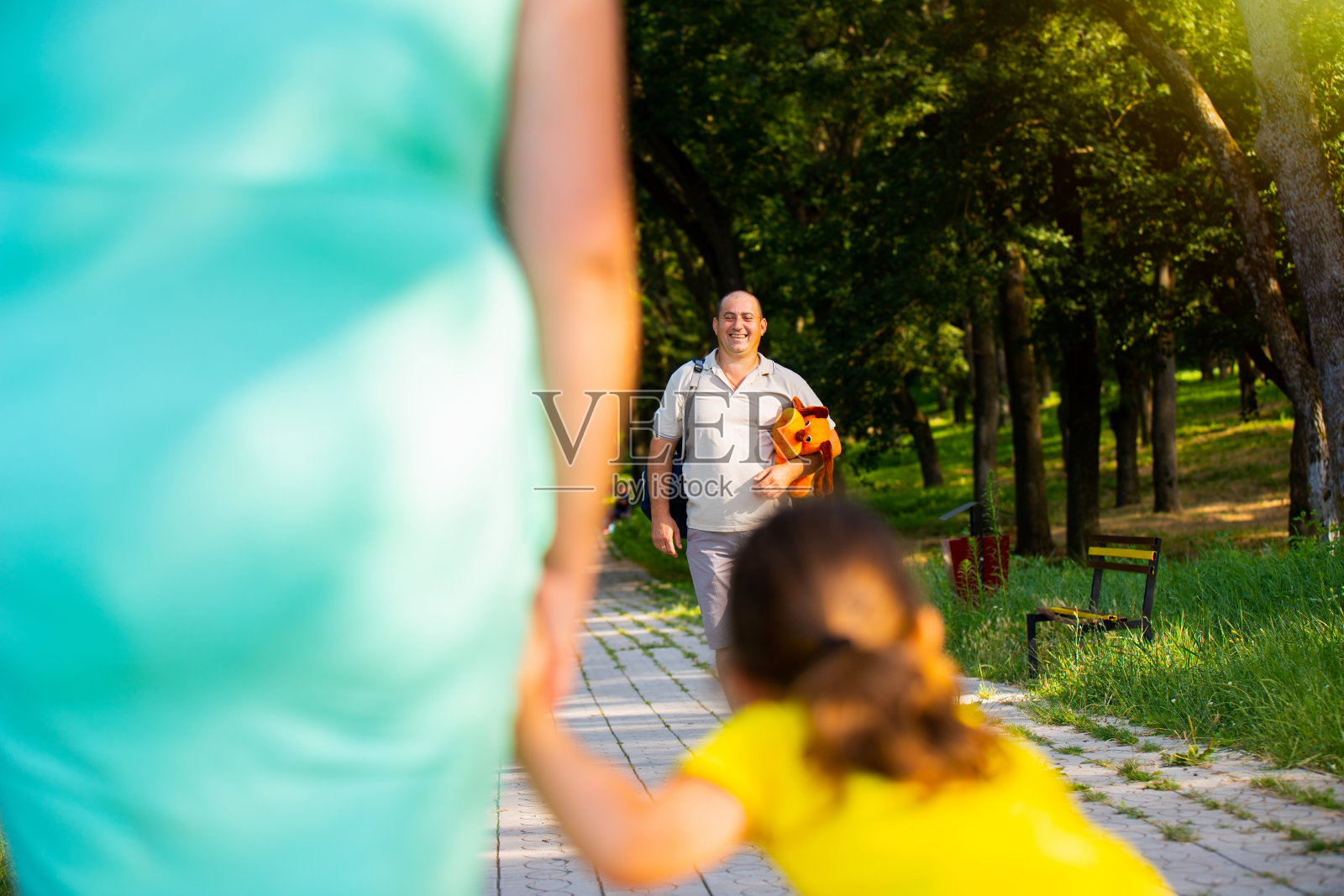 妈妈和小女孩的后视图照片等不及她的爸爸一起在公园度过洞日,他准备迎接他们,手里拿着一个柔软的狗玩具给他的女儿。照片摄影图片