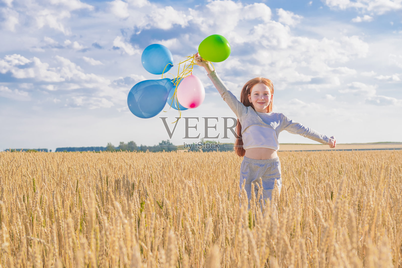 快乐的红发小女孩在麦田里奔跑,手里拿着彩色的气球,背景是蓝色的天空。照片摄影图片