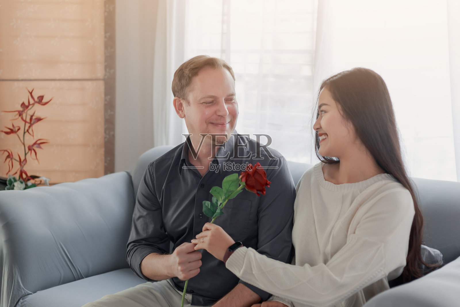 一对可爱的情侣在情人节用玫瑰摆出全家福的姿势,开心地笑着,关系很好。跨种族夫妇亚洲和高加索放松坐在沙发上在家里。照片摄影图片