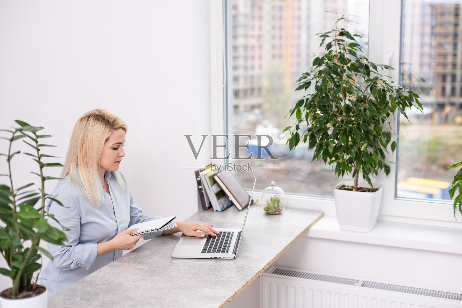 年轻的金发白种人女性摄影师坐在笔记本电脑木桌大窗户和灰色的墙工作照片摄影图片