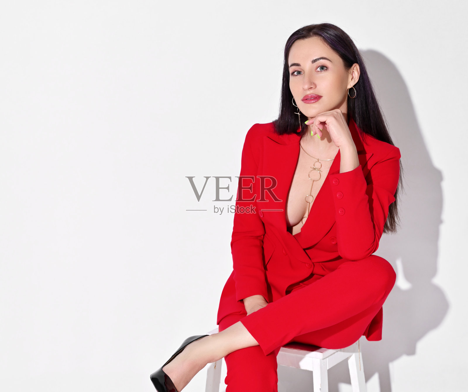年轻的黑发女人的肖像在红色的官方长裤套装,夹克与深领口赤裸的身体坐在凳子上照片摄影图片