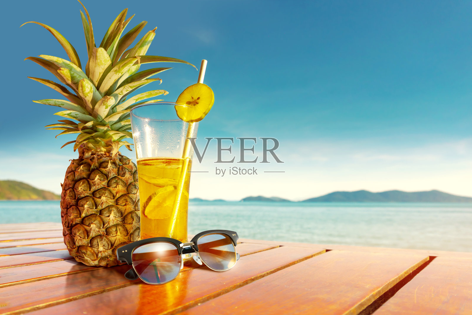 太阳眼镜和用菠萝和柠檬制成的果汁被放在海边的木桌上。照片摄影图片
