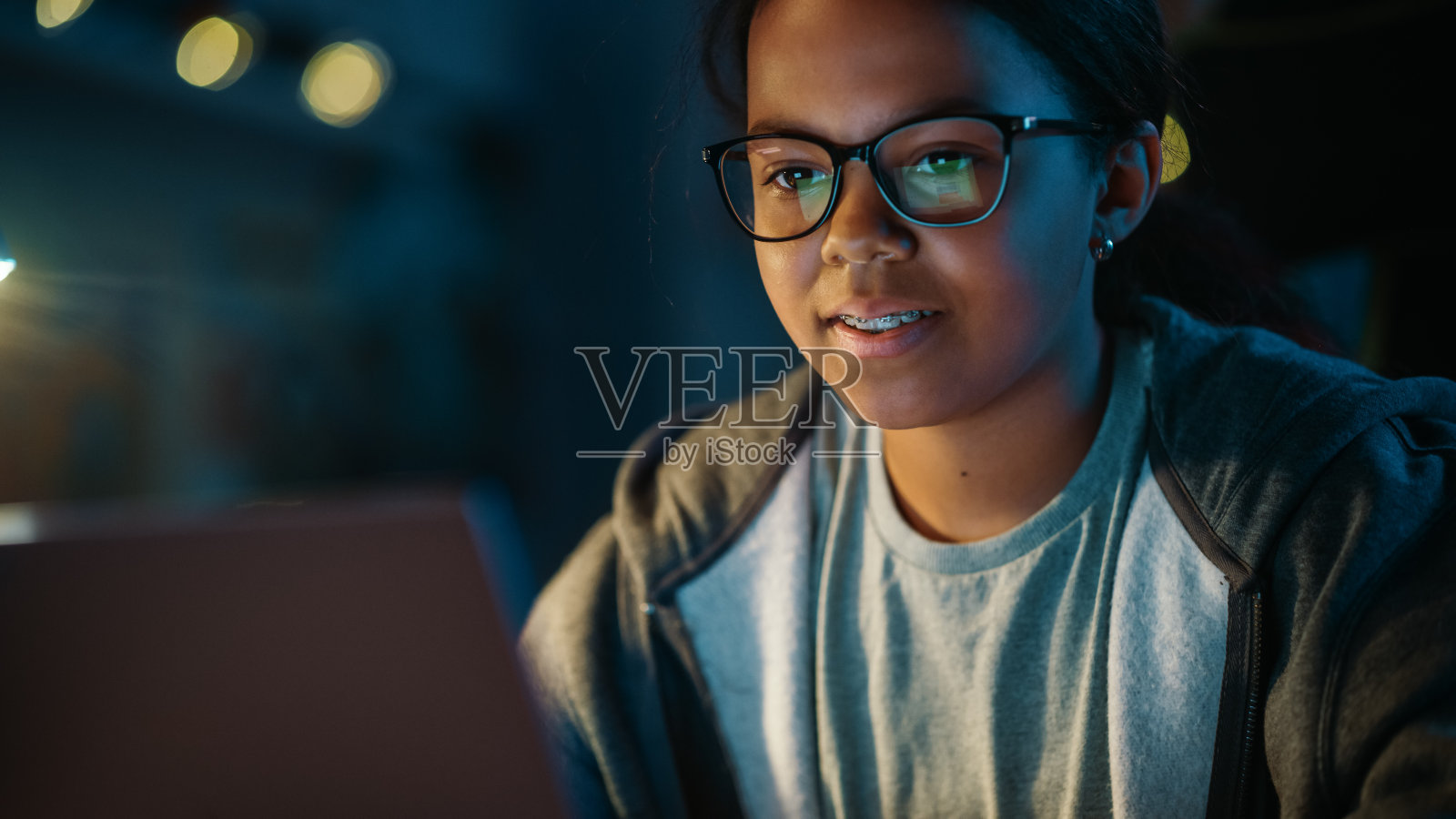 十几岁的多种族黑人女孩在家里一个黑暗舒适的房间里使用笔记本电脑。她正在浏览在线教育研究。学习科学学校作业观念。照片摄影图片