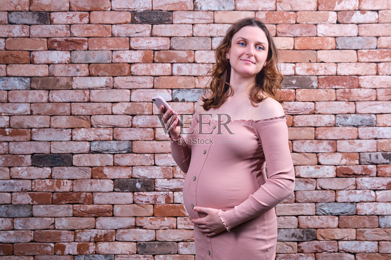 快乐孕妇身穿红色连衣裙,手机贴在砖墙背景上照片摄影图片