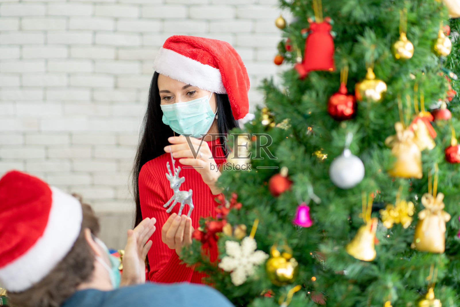 在新冠疫情期间,一名戴口罩的白人女子和他的夫妇在他家装饰圣诞树,庆祝节日照片摄影图片