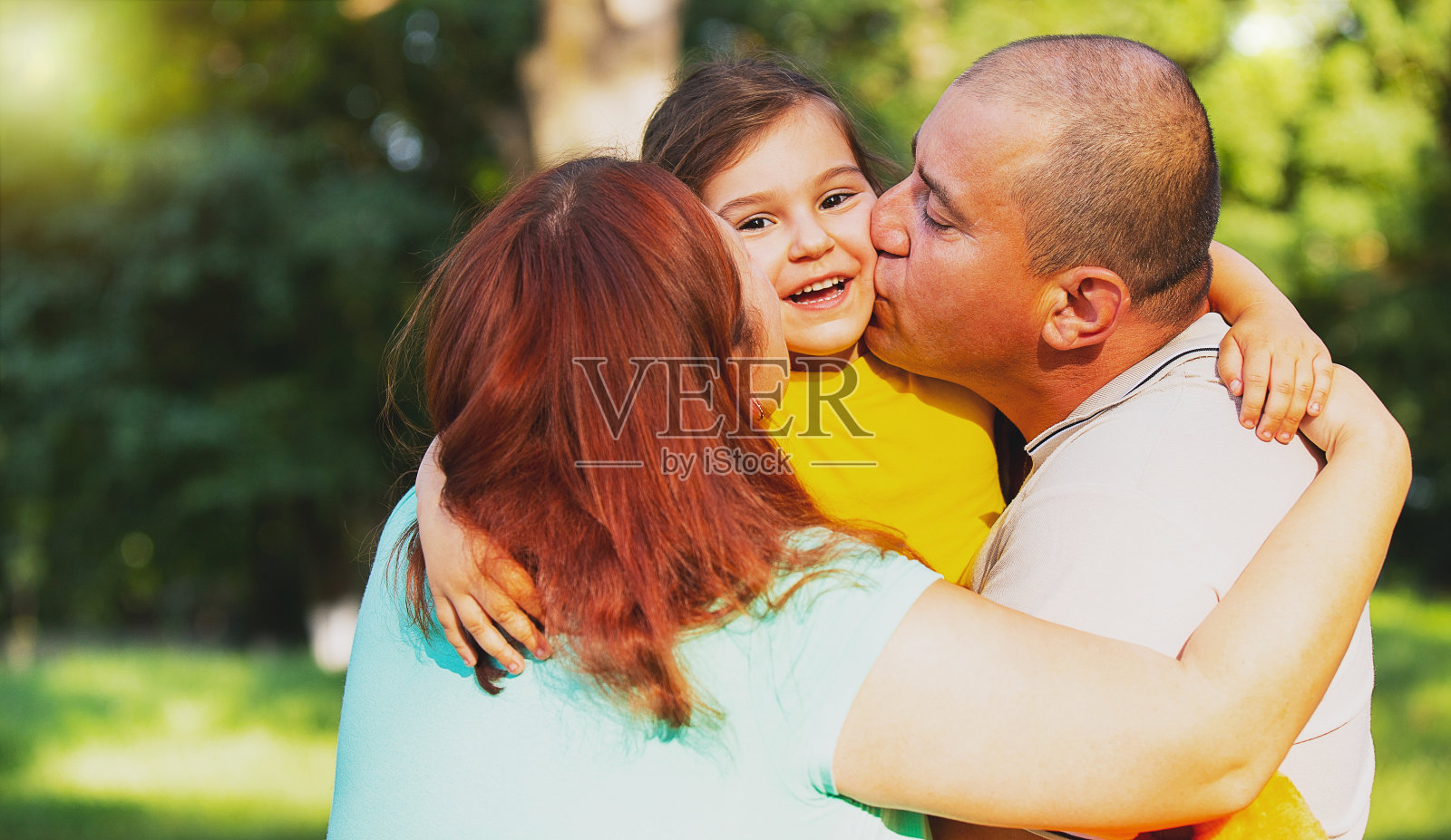 年轻微笑的家庭和孩子在一起玩,爱的夫妇抱着他们快乐的女儿在他们的手和亲吻她的脸颊。照片摄影图片