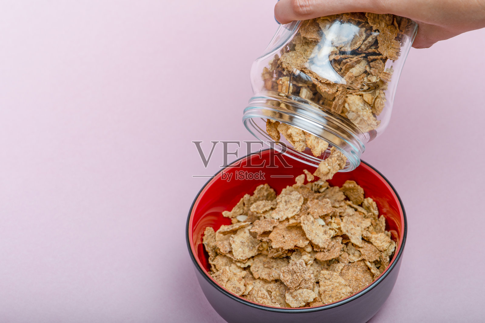 粉红色背景上的什锦麦片。用手将干麦片片倒入碗中。当用麦片做麦片时,把麦片从玻璃罐里倒进黑色的碗里照片摄影图片