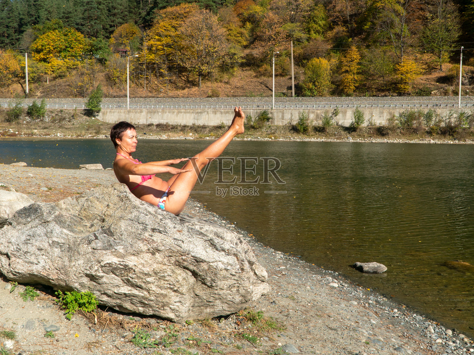秋天,一个身穿比基尼的女子在湖边的一块大石头上做着“纳佛式”照片摄影图片