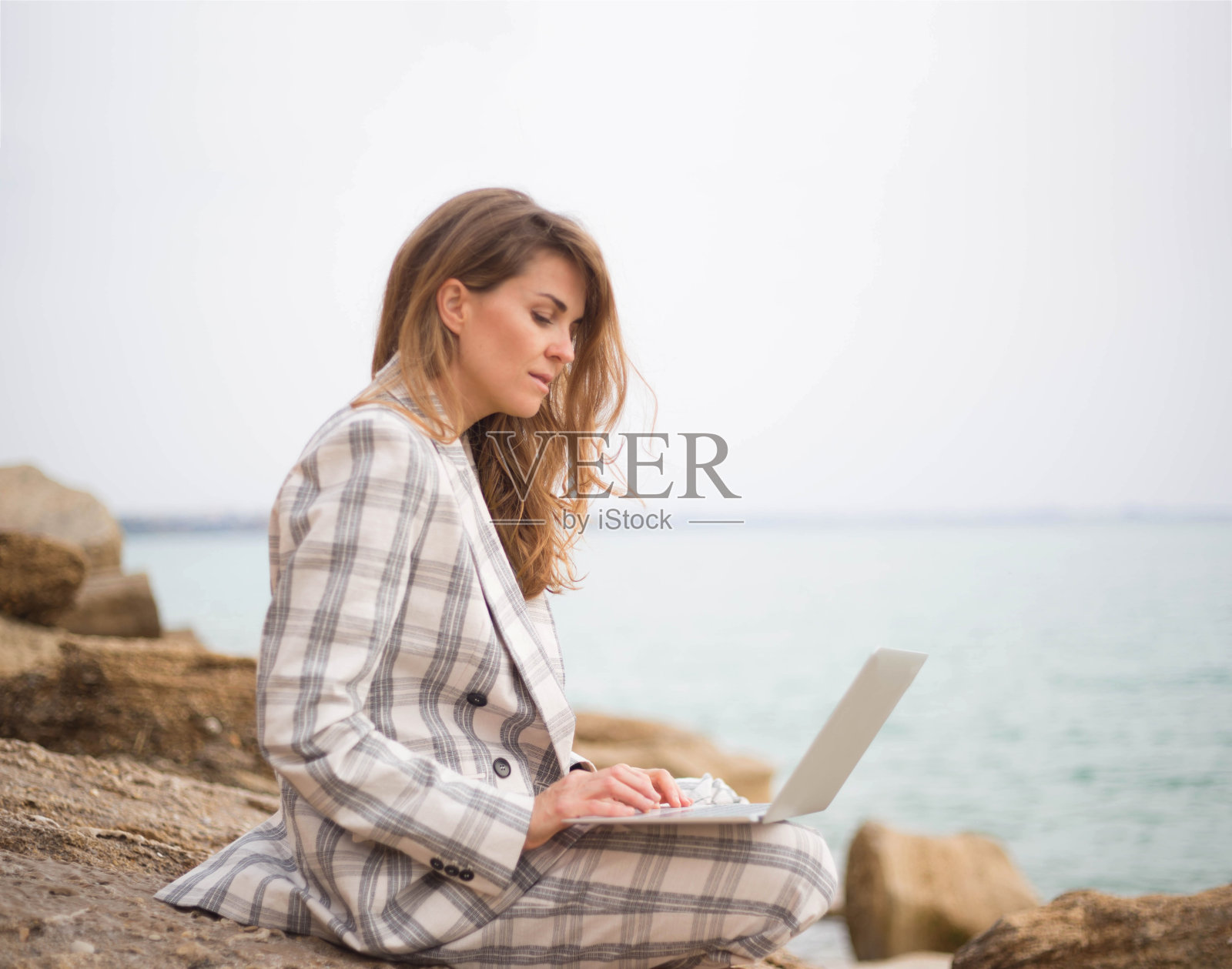 一位年轻女子坐在海边的一块石头上,手里拿着一台笔记本电脑。远程工作的概念照片摄影图片