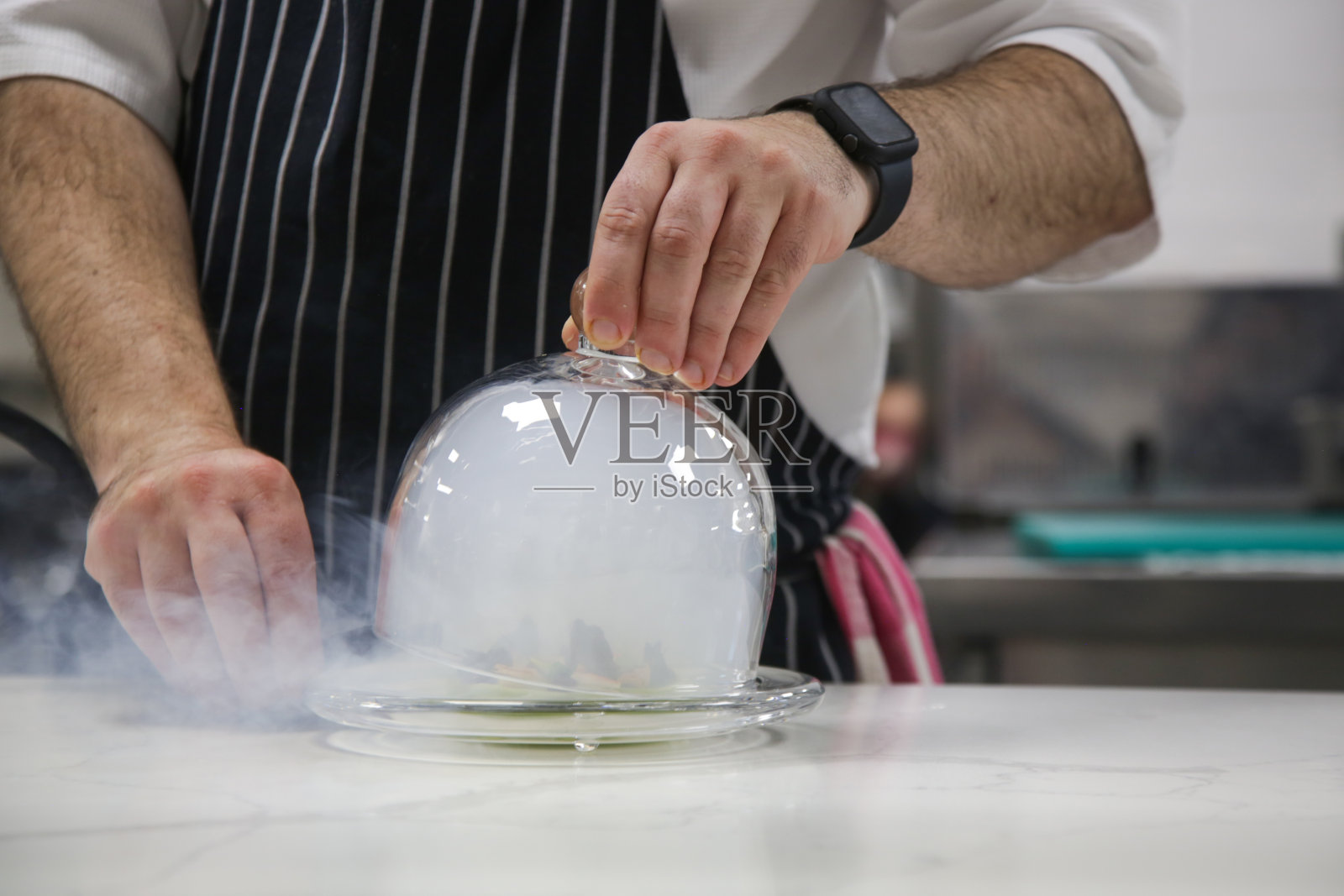 厨师的手从盛着热食物的盘子里举起玻璃钟形罩,餐厅里烟雾缭绕。精致的菜肴,创意的餐厅用餐理念,高级定制食物。照片摄影图片