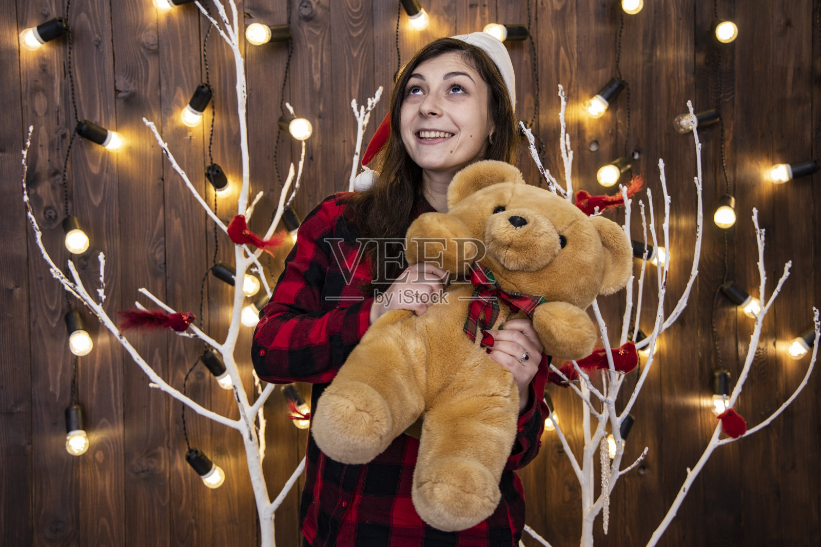 快乐可爱迷人的少女,圣诞风格的肖像拥抱泰迪熊玩具,舒适的室内装饰,用花环灯温暖的黄色照明照明背景照片摄影图片