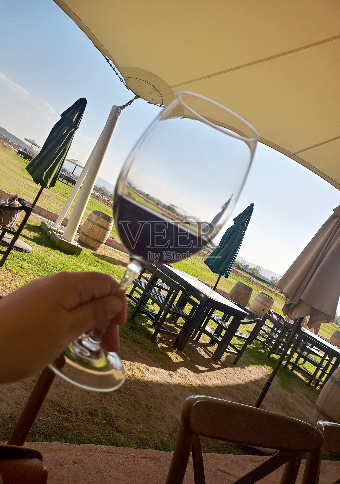 在前景中享用一杯红酒,后面是户外木制野餐桌、雨伞和酒桶照片摄影图片